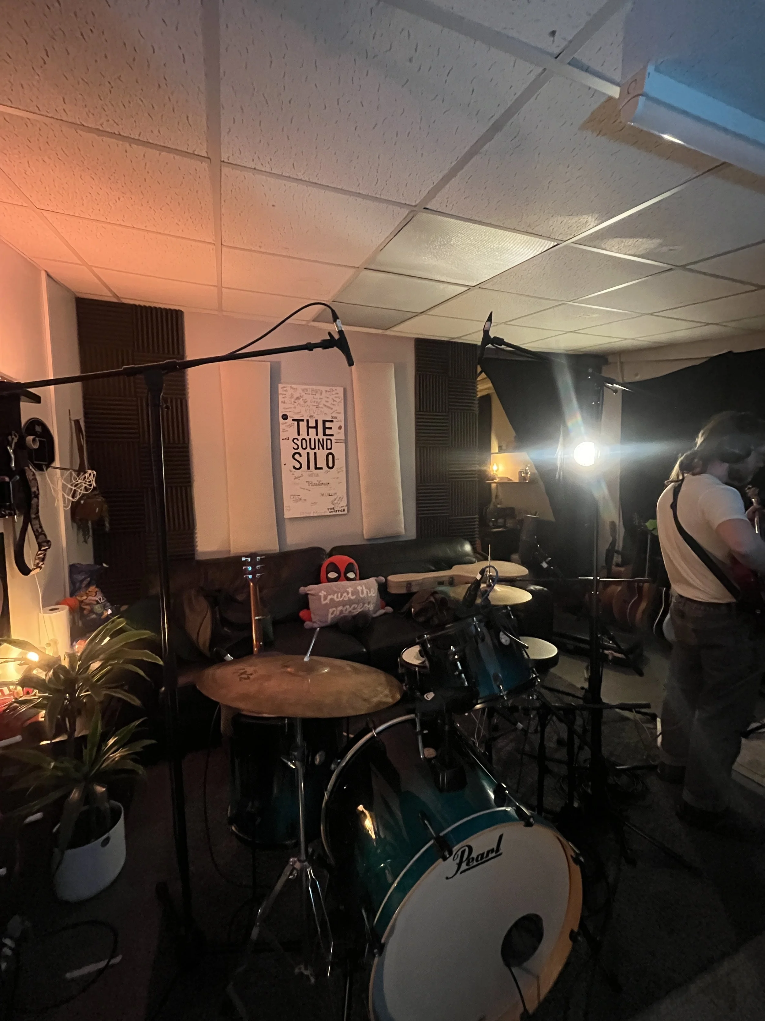 Music recording studio with drums, microphones, and a person tuning a guitar. Soundproofing foam on walls, a poster on the wall reads "The Sound Silo," and there is dim lighting with stage lights on.