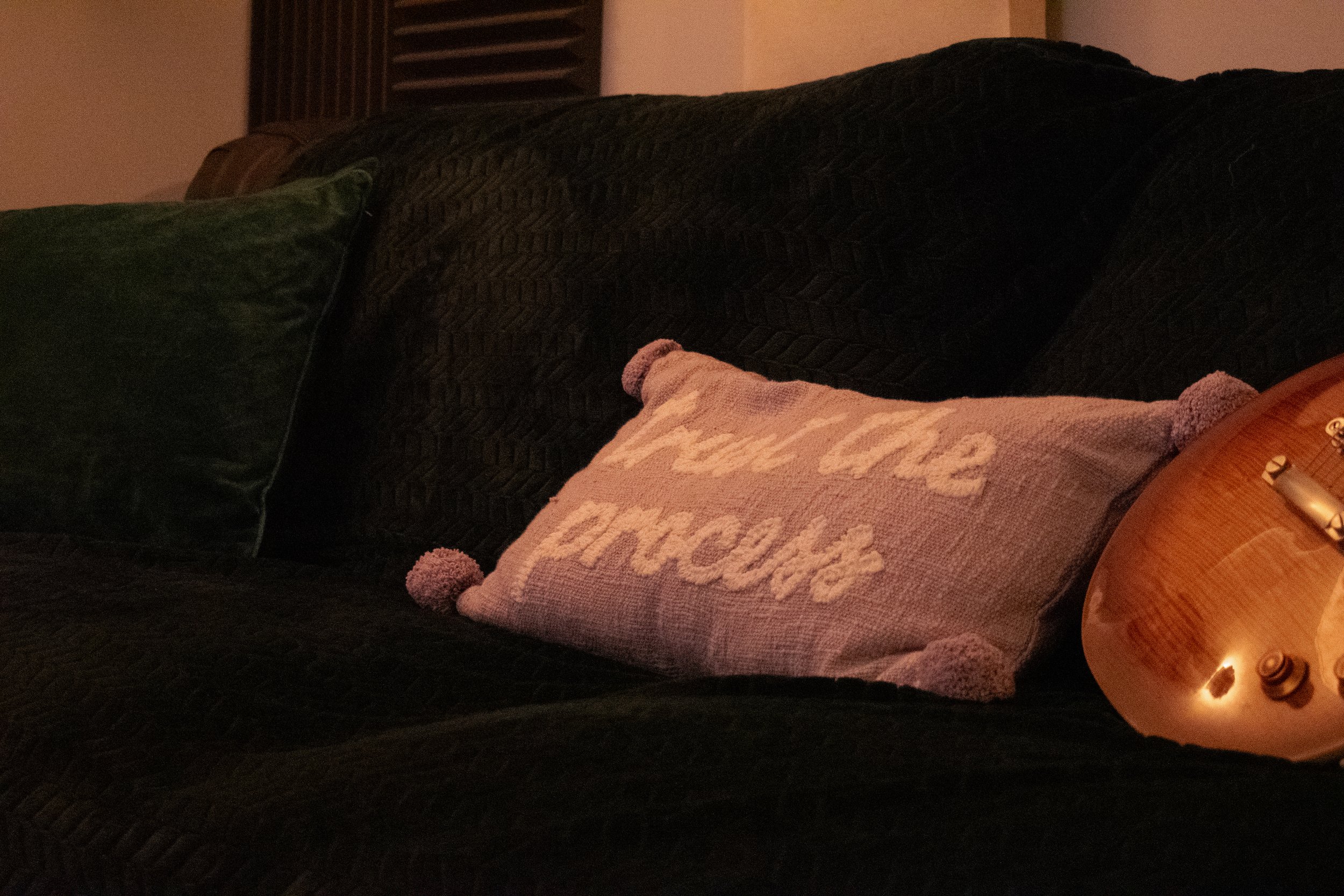 Close-up of a dark-colored textured couch with a green pillow, a pink decorative pillow that says 'You Are Amazing' with pom-pom edges, and part of a wooden stringed instrument resting on the arm of the couch.