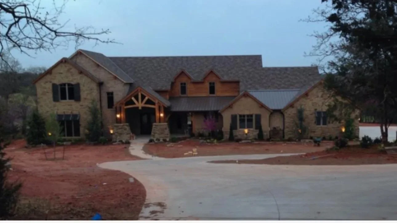 A large house with stone exterior and multiple roof lines, situated on a curved driveway with a landscaped yard.