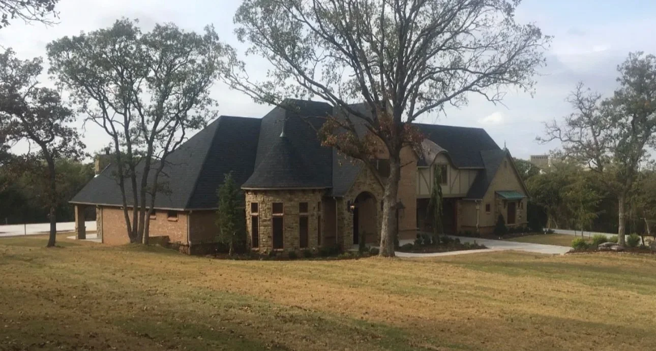 Front view of a large brick house with a black roof, surrounded by a lawn and several trees.