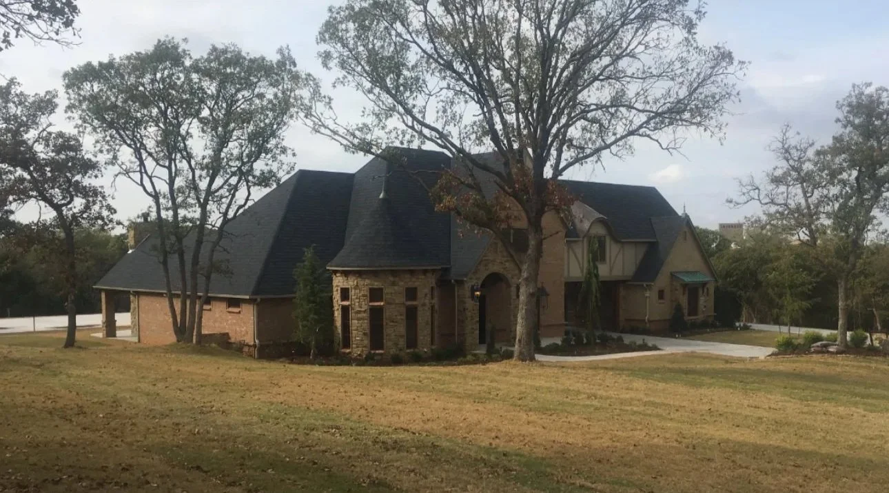 Large house with a brick and stone exterior, multiple sections, and a dark roof, surrounded by several trees on a spacious lawn.