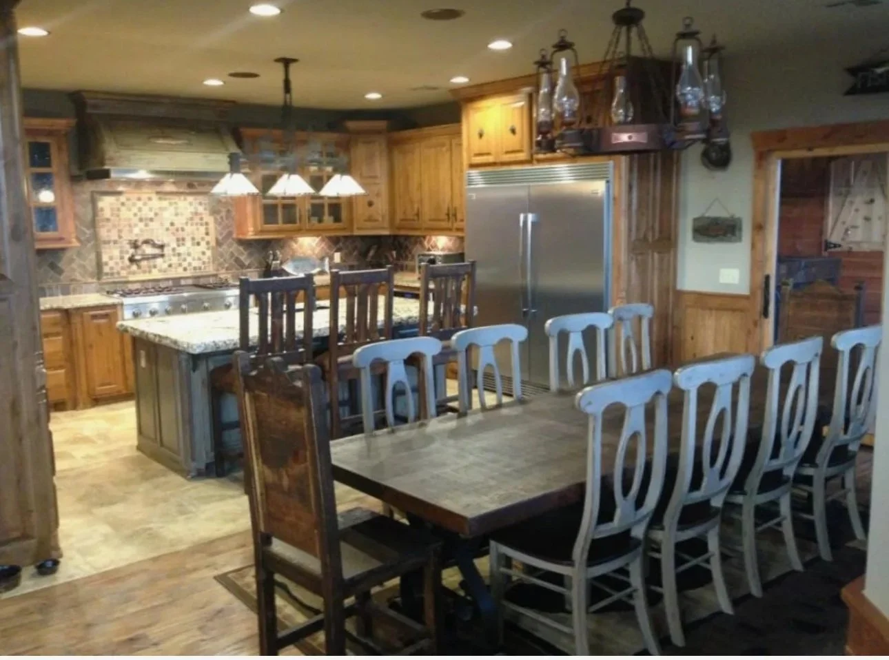 This image shows a rustic-style kitchen and dining area with wooden cabinets, a large dining table with multiple chairs, a kitchen island, and hanging lantern lighting.