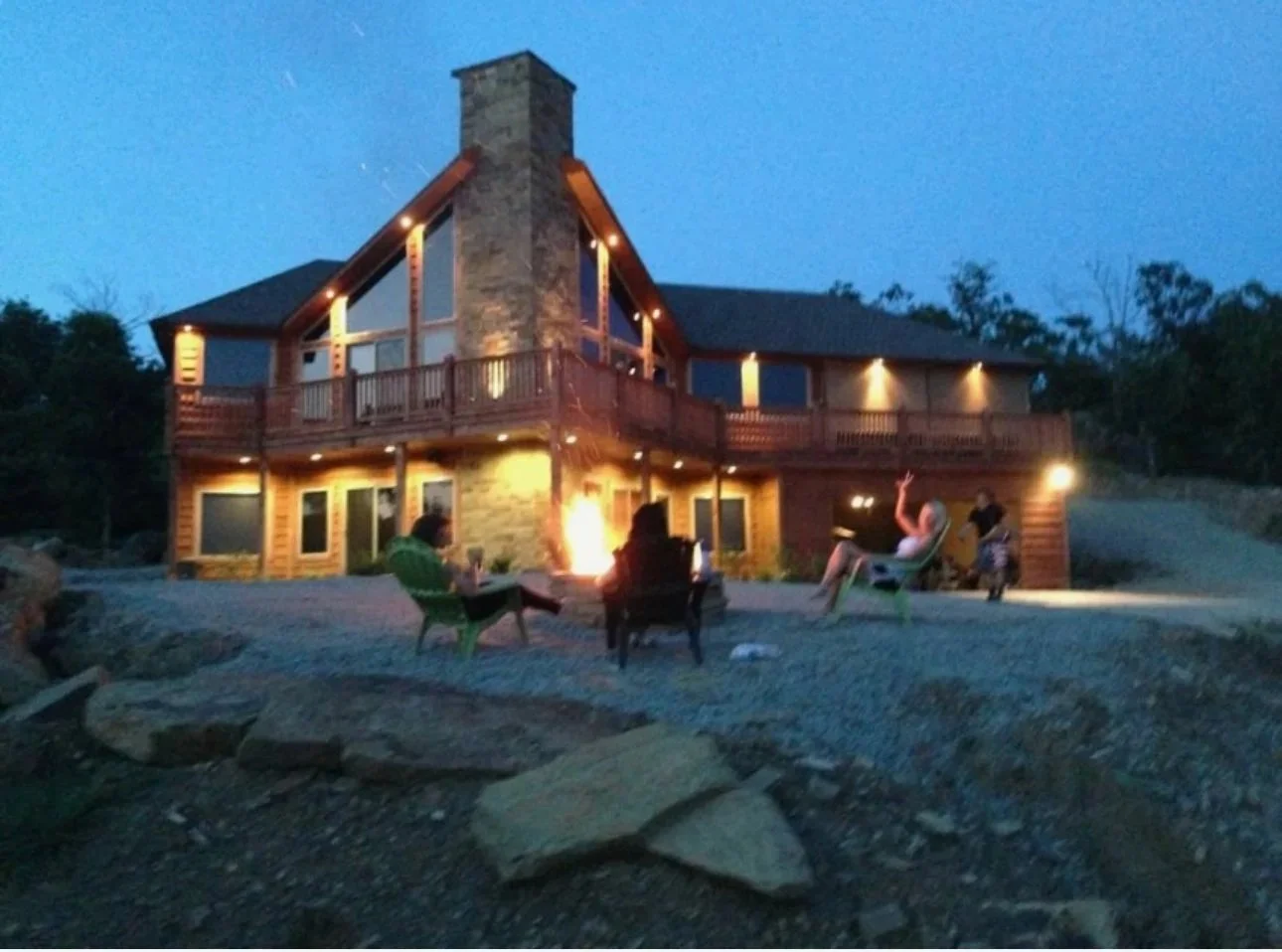 A group of people sitting outside a two-story house at dusk, with warm lights illuminating the exterior and a chimney, surrounded by trees and rocks.