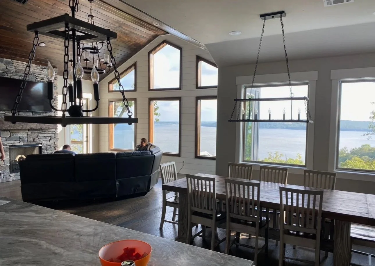 Living room with large windows overlooking a lake, featuring a stone fireplace, a sofa with two people, and a wooden dining table with chairs.
