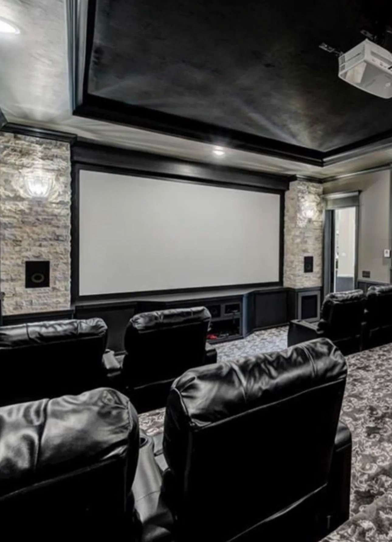 Home theater room with large projection screen, black leather recliner seats, stone accent walls, and ceiling-mounted projector.