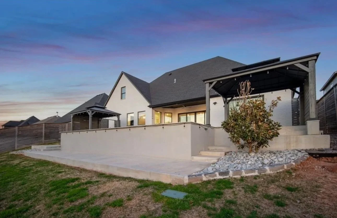 Backyard view of a modern house with a large concrete patio, two covered pergolas, and a small landscaped area with a shrub and rocks. Fencing surrounds the yard, with a clear evening sky overhead.