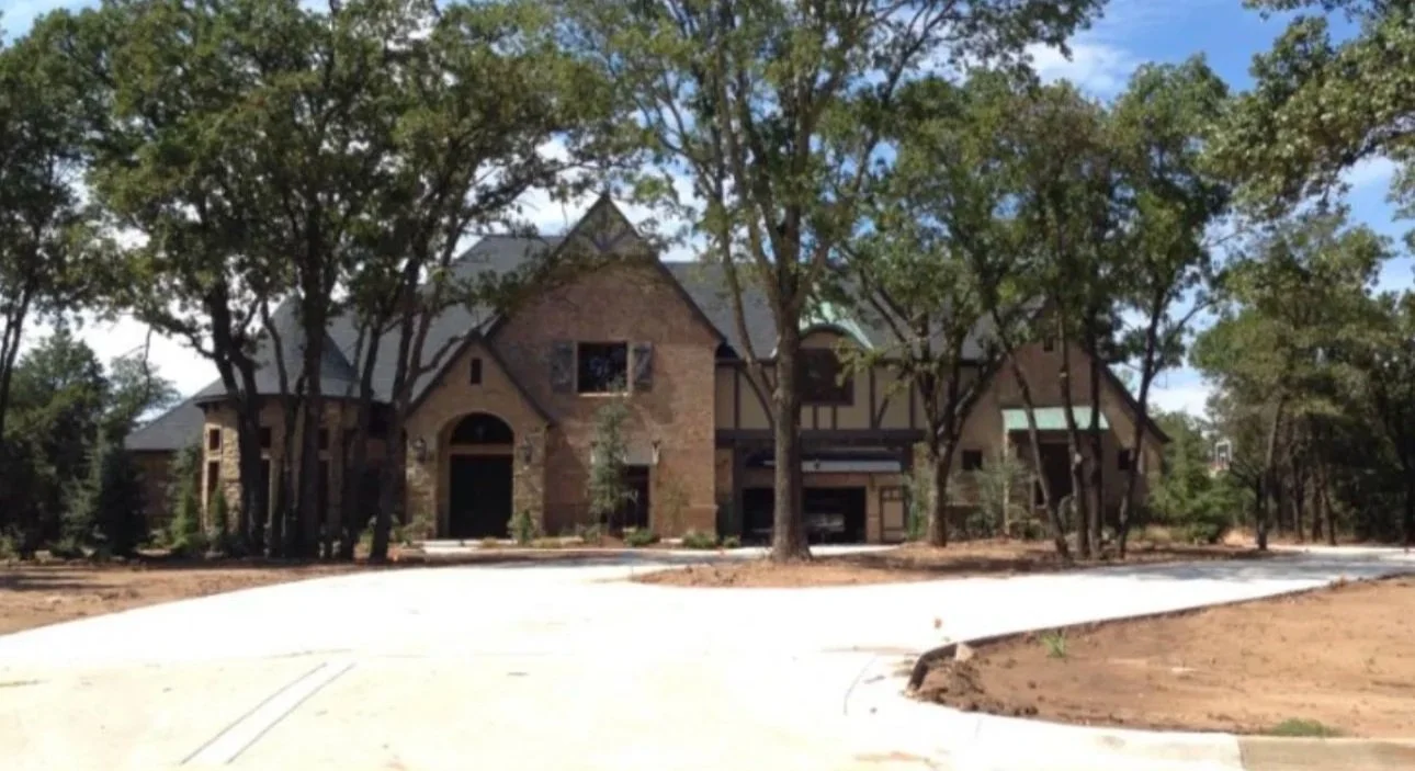 Large brick and wood house surrounded by trees with a concrete driveway in front.
