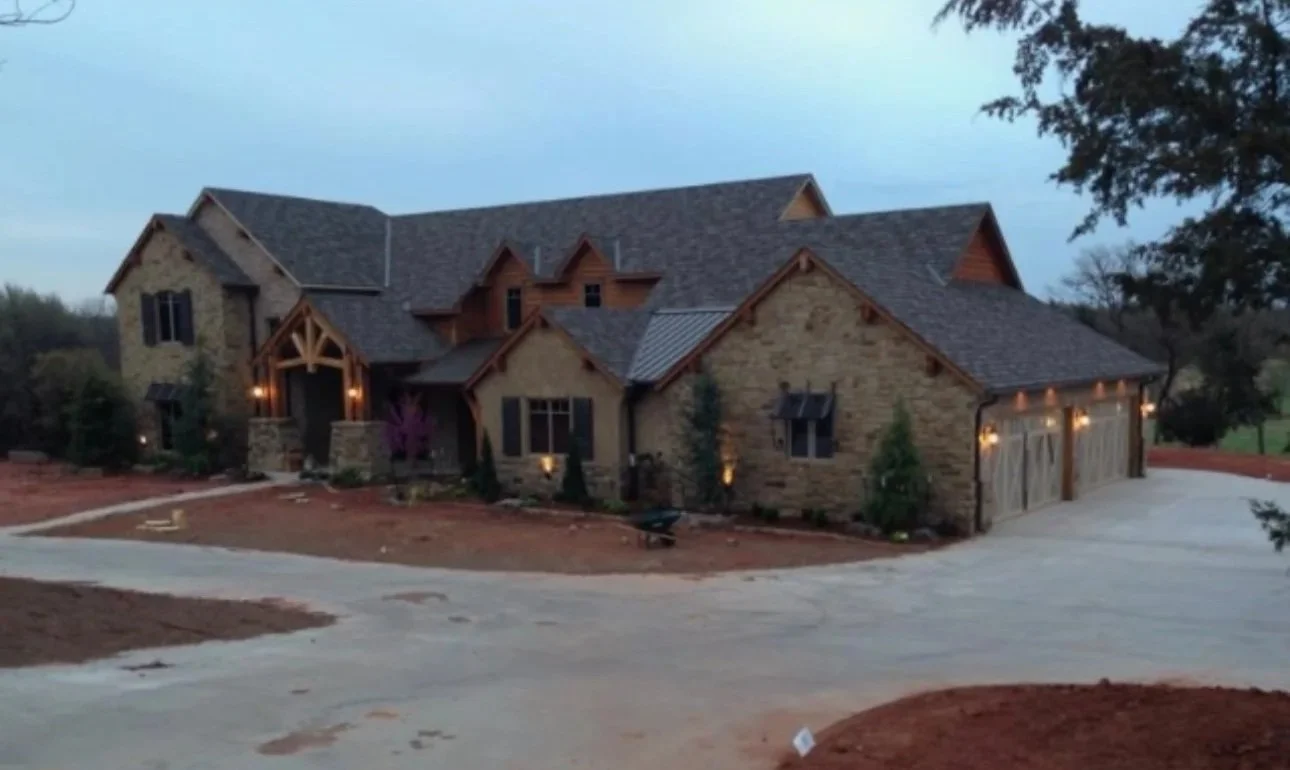 Large house with stone walls, wooden accents, multiple gabled roofs, and a three-car garage, surrounded by a partially landscaped yard with trees and dirt patches.