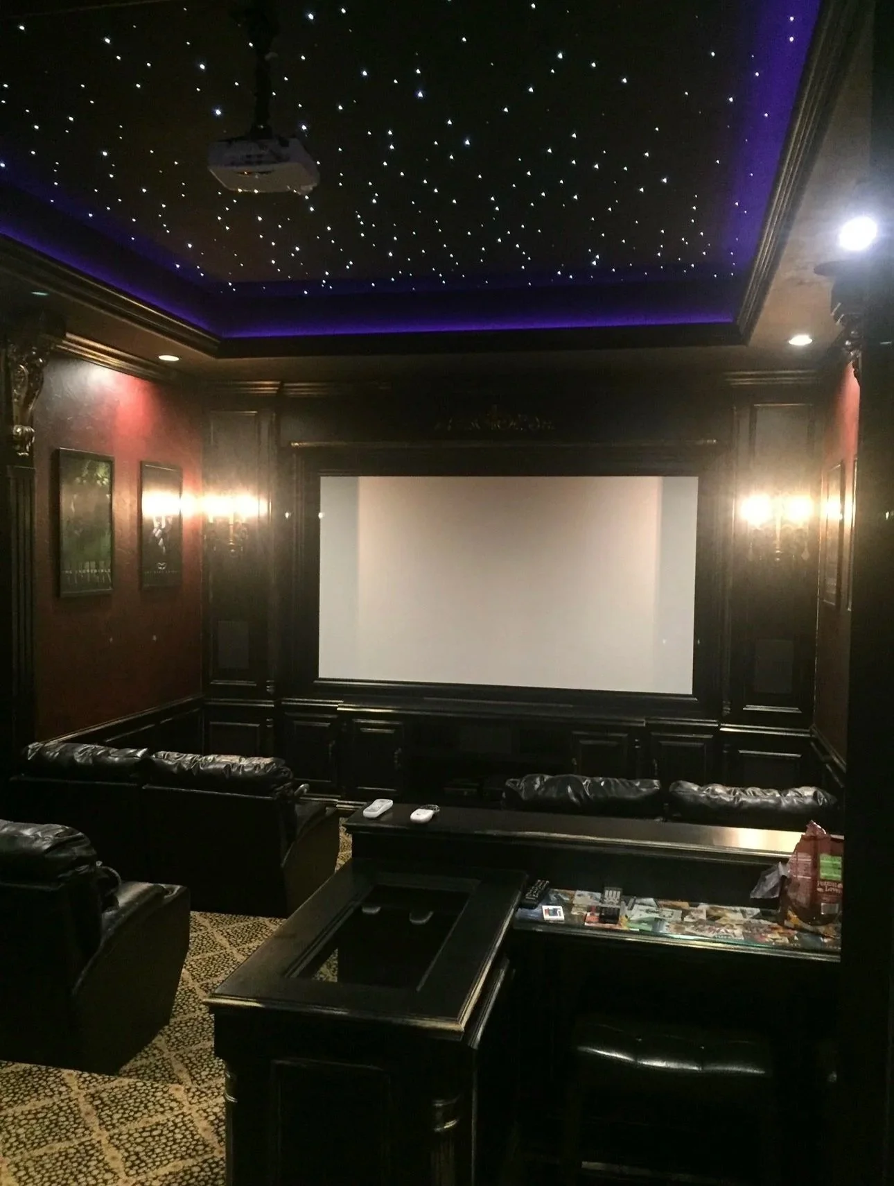 A luxurious home theater room with a large projection screen, leather sofas, a dark wood coffee table, and a starry ceiling with LED lights. There are wall-mounted lights and movie posters on the walls.