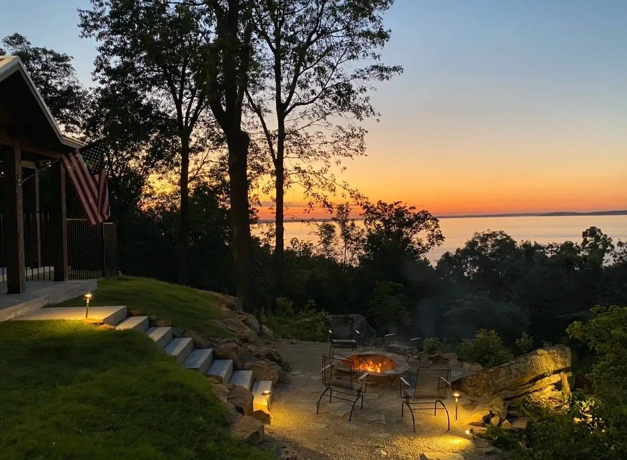 A backyard patio with a fire pit surrounded by chairs, overlooking a river at sunset. The patio has steps leading up to a house with an American flag, and the scene is illuminated by small outdoor lights.