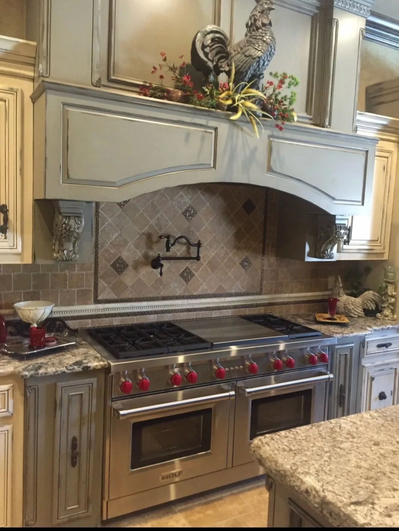A kitchen with a built-in stainless steel Wolf stove with two oven doors and six red control knobs, beige cabinetry with ornate detailing, and decorative rooster and hen sculptures with floral arrangements on the range hood mantle.