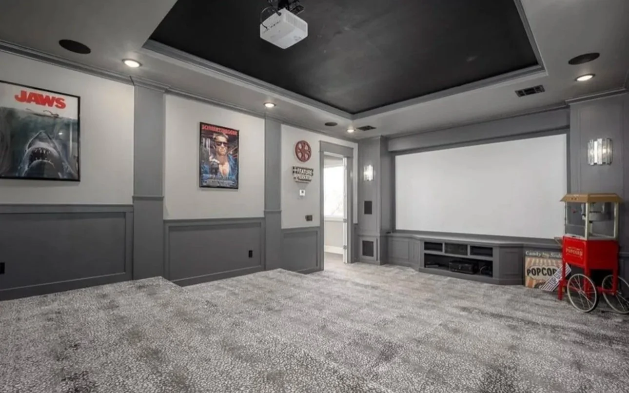 Empty home theater room with gray walls, black ceiling, movie posters on the wall, popcorn and movie memorabilia, and a large blank screen.
