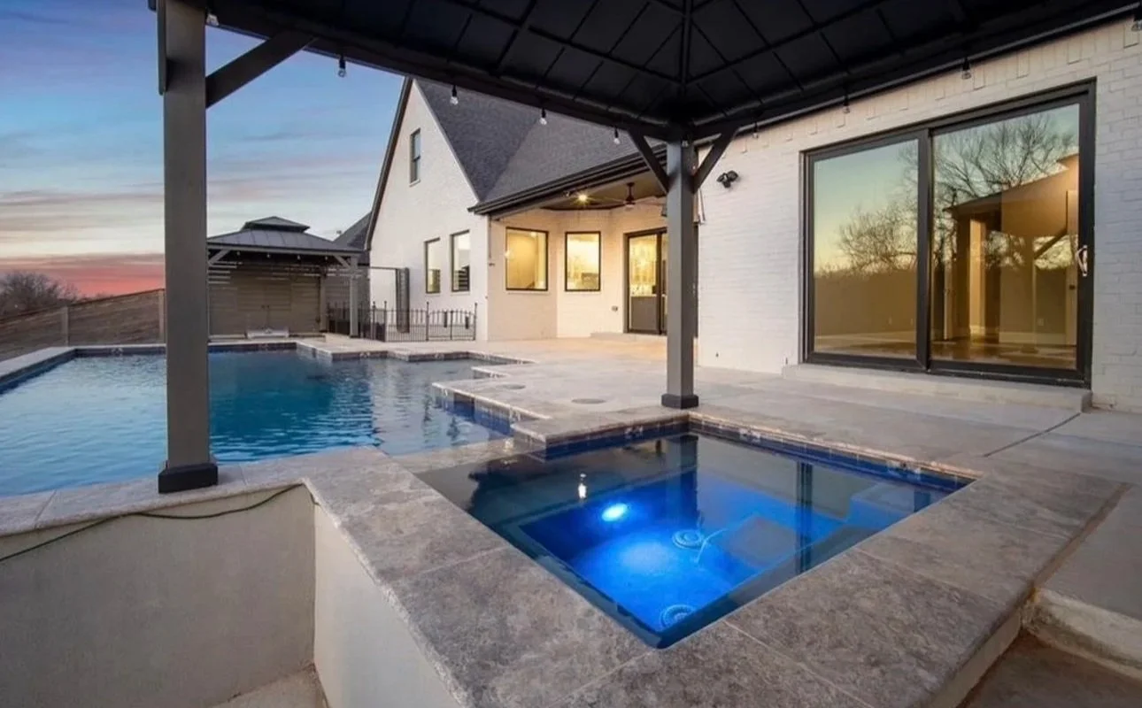Modern house patio with a swimming pool and hot tub, under a covered patio area at sunset, with large windows reflecting trees outside.