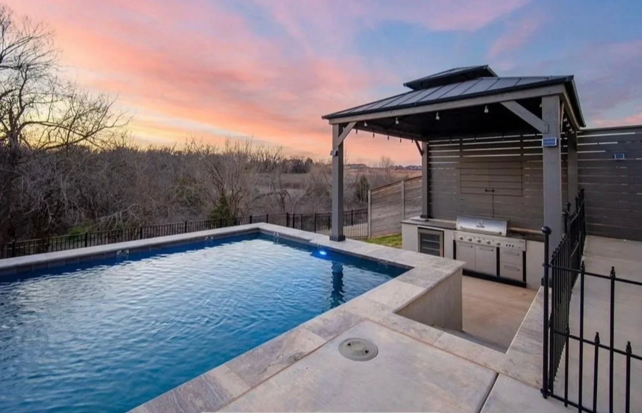 An outdoor backyard space featuring a swimming pool with a spa, a covered outdoor kitchen with a grill, and a sunset sky with pink and blue hues.