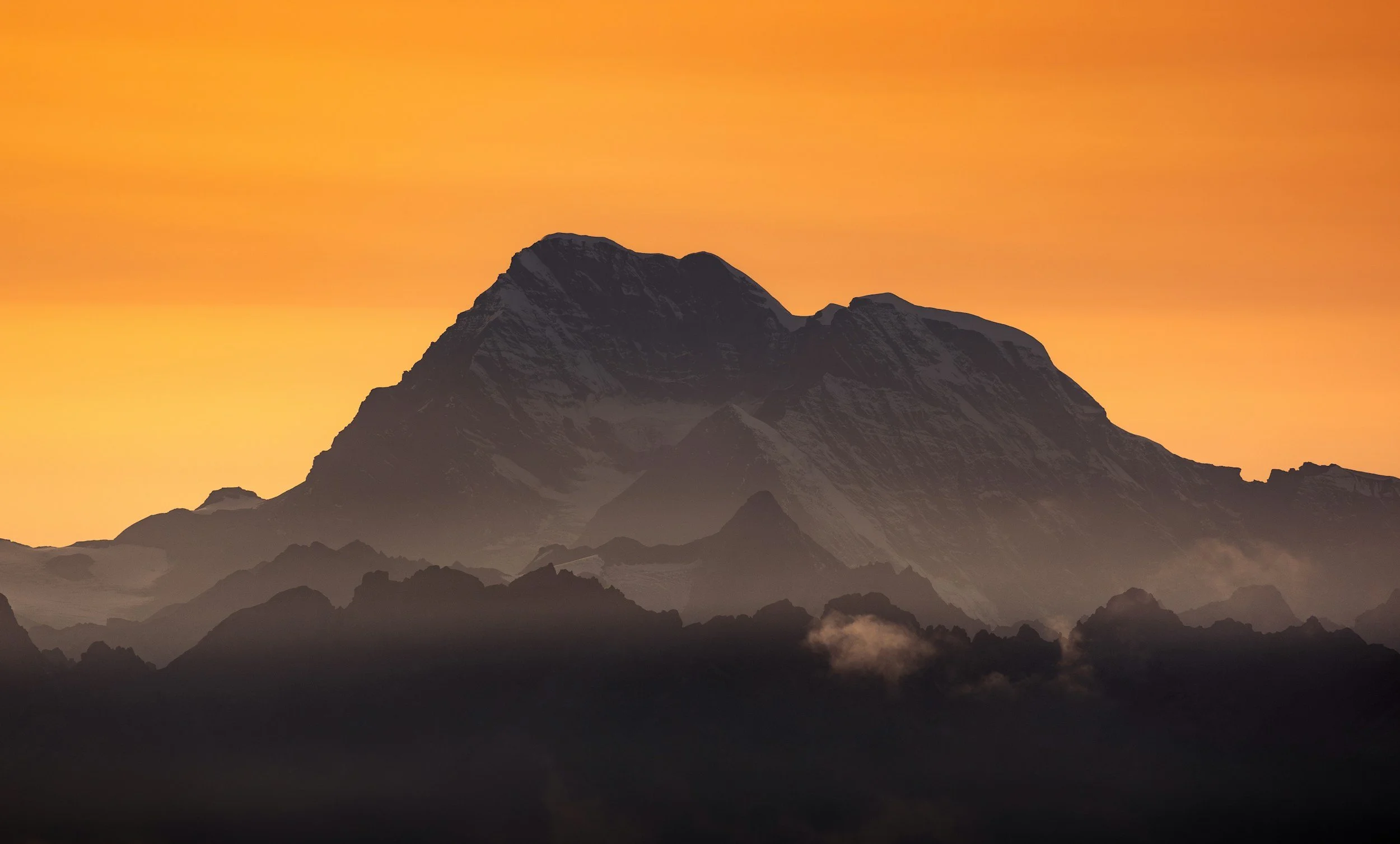 A mountain during sunset with an orange sky, dark silhouette of peaks, and some clouds around the mountain base.