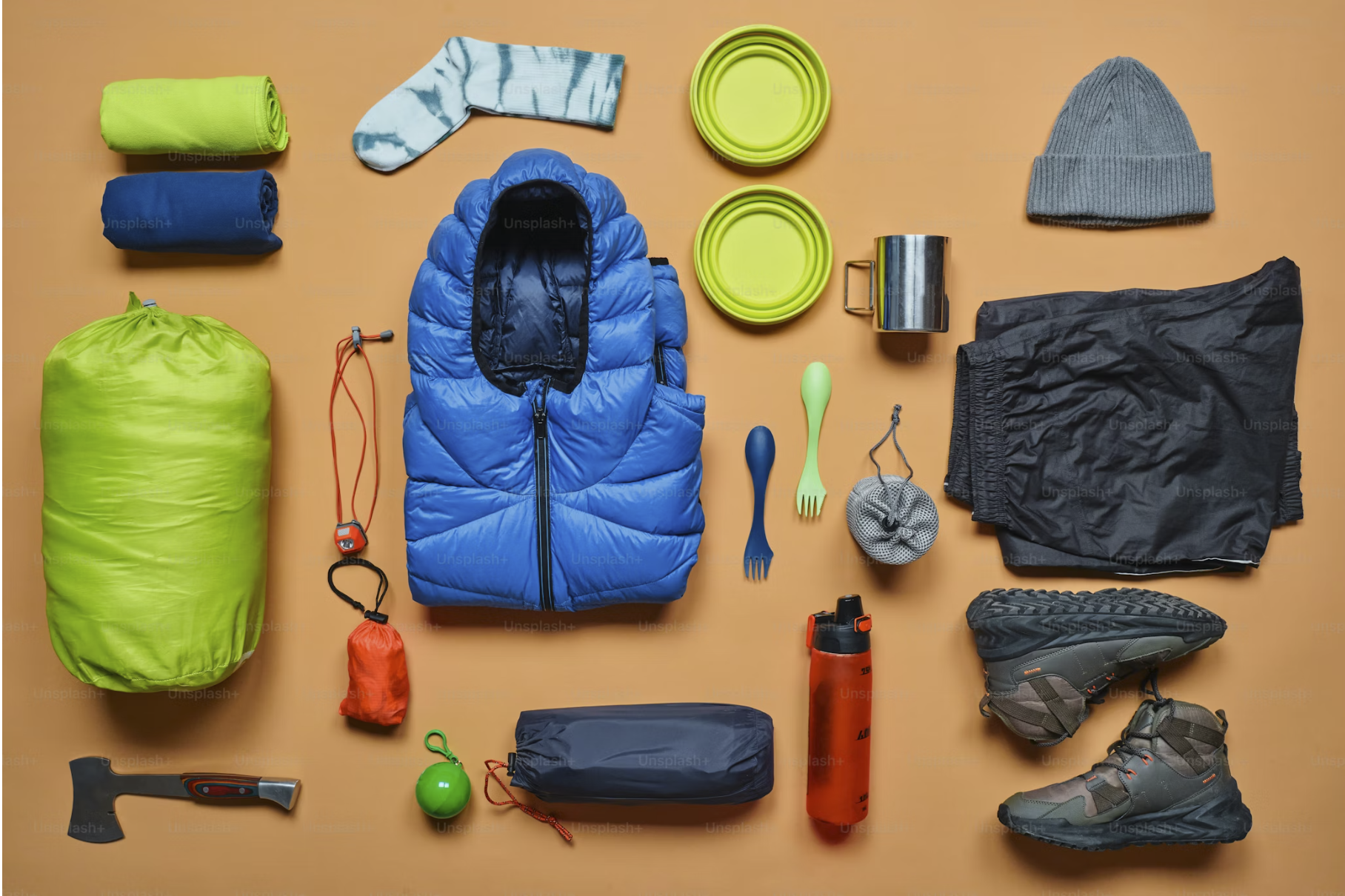 Camping gear laid out on an orange background including a green sleeping bag, a blue puffer jacket, a gray knit hat, black shorts, hiking boots, a green ball, a little axe, a green bowl, a yellow plate, a green spork and fork, a metal mug, a red water bottle, a gray pack, a small gray pouch, a blue roll of tape, socks, a small orange bag, a green carabiner, and an orange whistle.