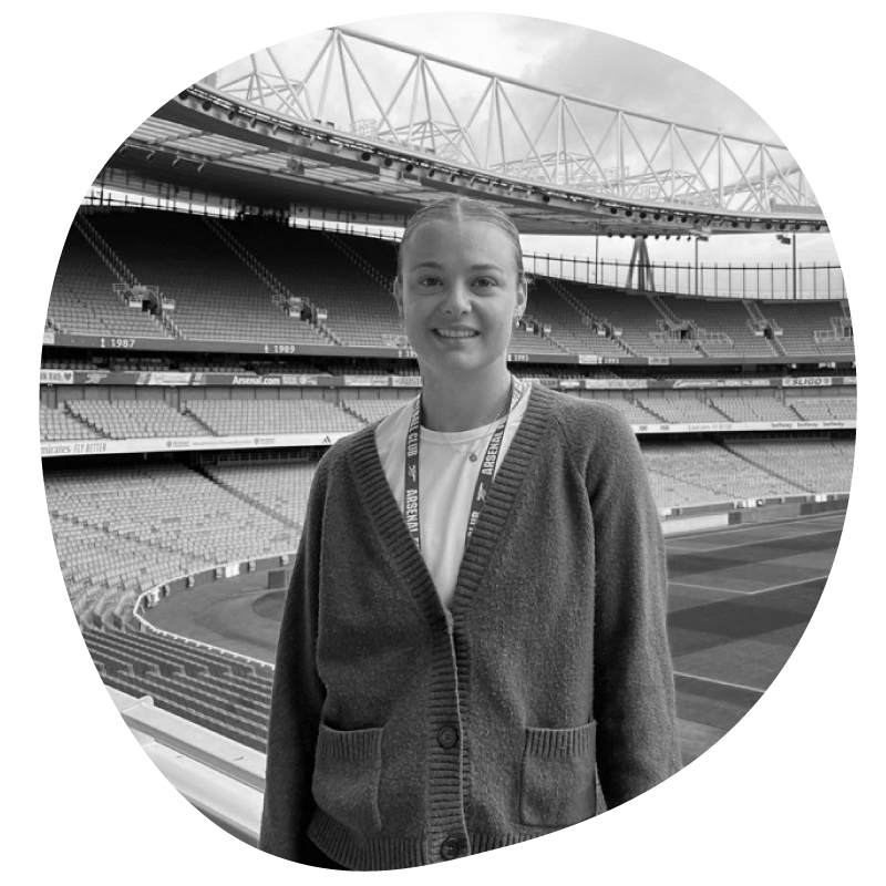 A young girl standing in a sports stadium, smiling, with the field and empty seats in the background, in black and white.