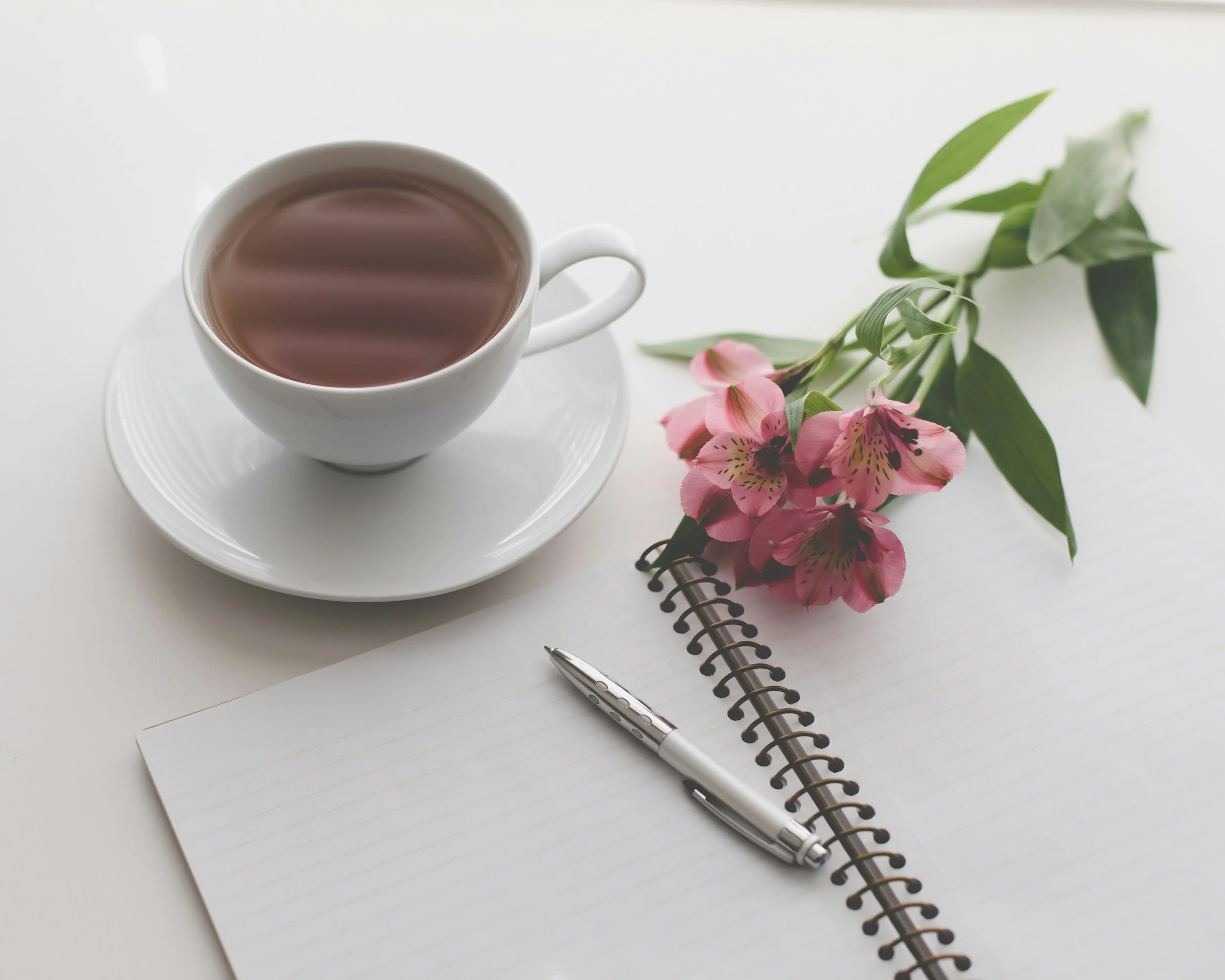 A white cup of tea or coffee on a white saucer, a pink flower with green leaves, a silver pen, and a lined spiral notebook on a white surface.