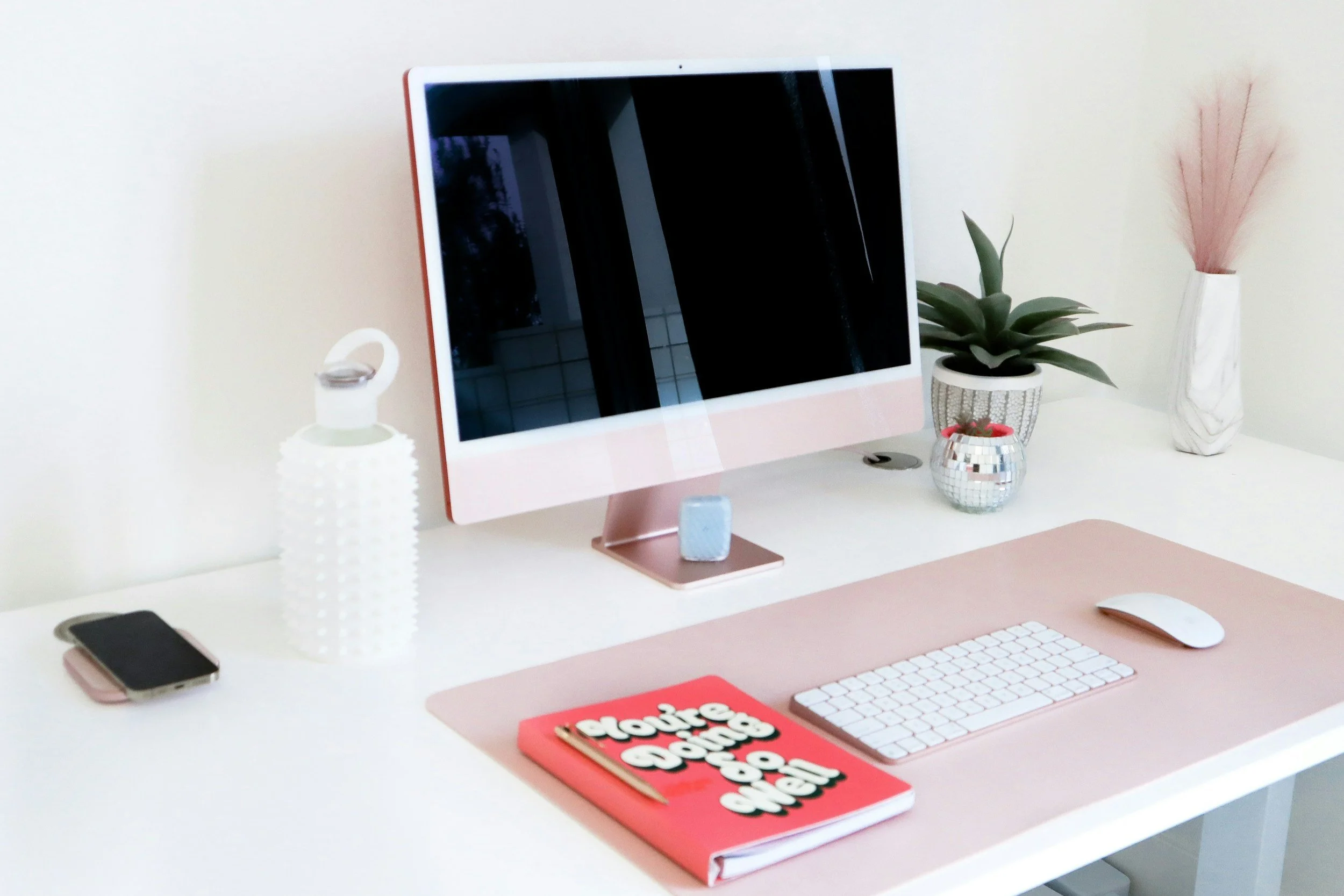 A white desk with a pink monitor, white keyboard, mouse, and pink desk mat. Items include a black smartphone, a white textured water bottle, a small blue cube, a red notebook with white text, a small disco ball, a potted succulent, and a white vase with pink pampas grass.