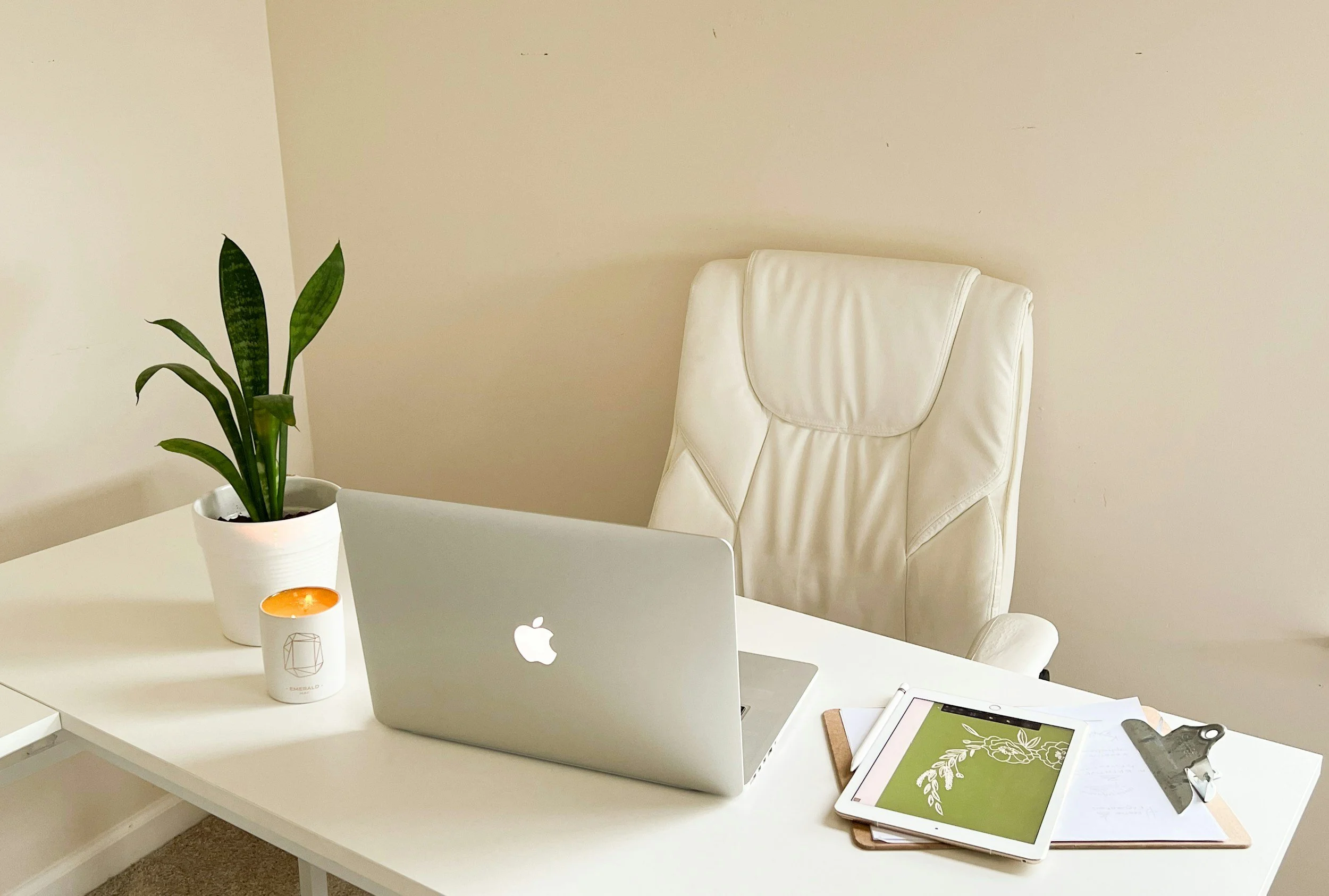 A white office desk with an open Apple MacBook, a white potted snake plant, a lit candle, a clipboard with papers, and a tablet with a green and white decorative screen. Behind the desk is a white leather office chair, and the background is a beige wall.