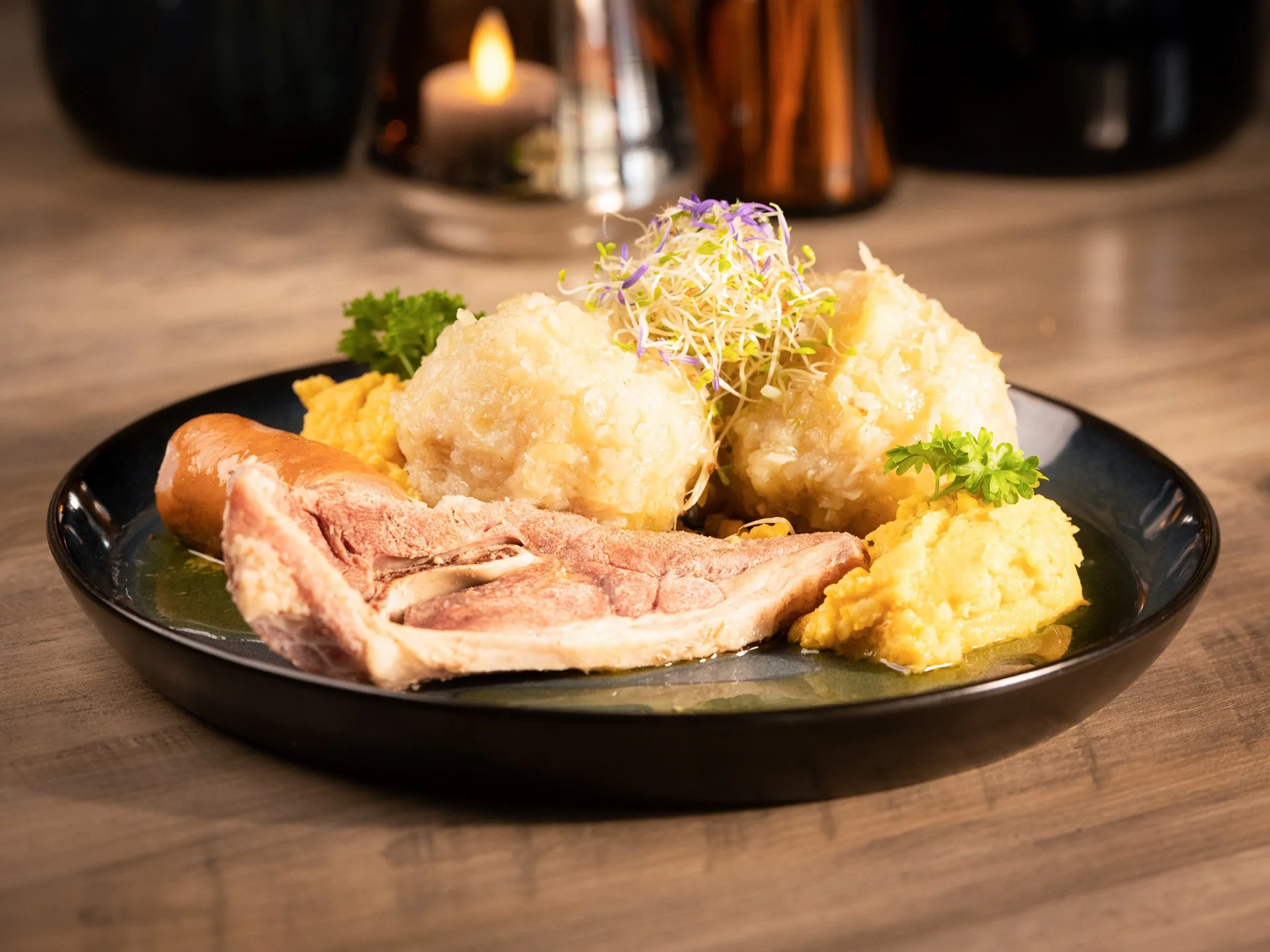 A plate of mashed potatoes with gravy, a slice of cooked lamb, mashed sweet potatoes, and garnishes of parsley and microgreens, served on a black plate.