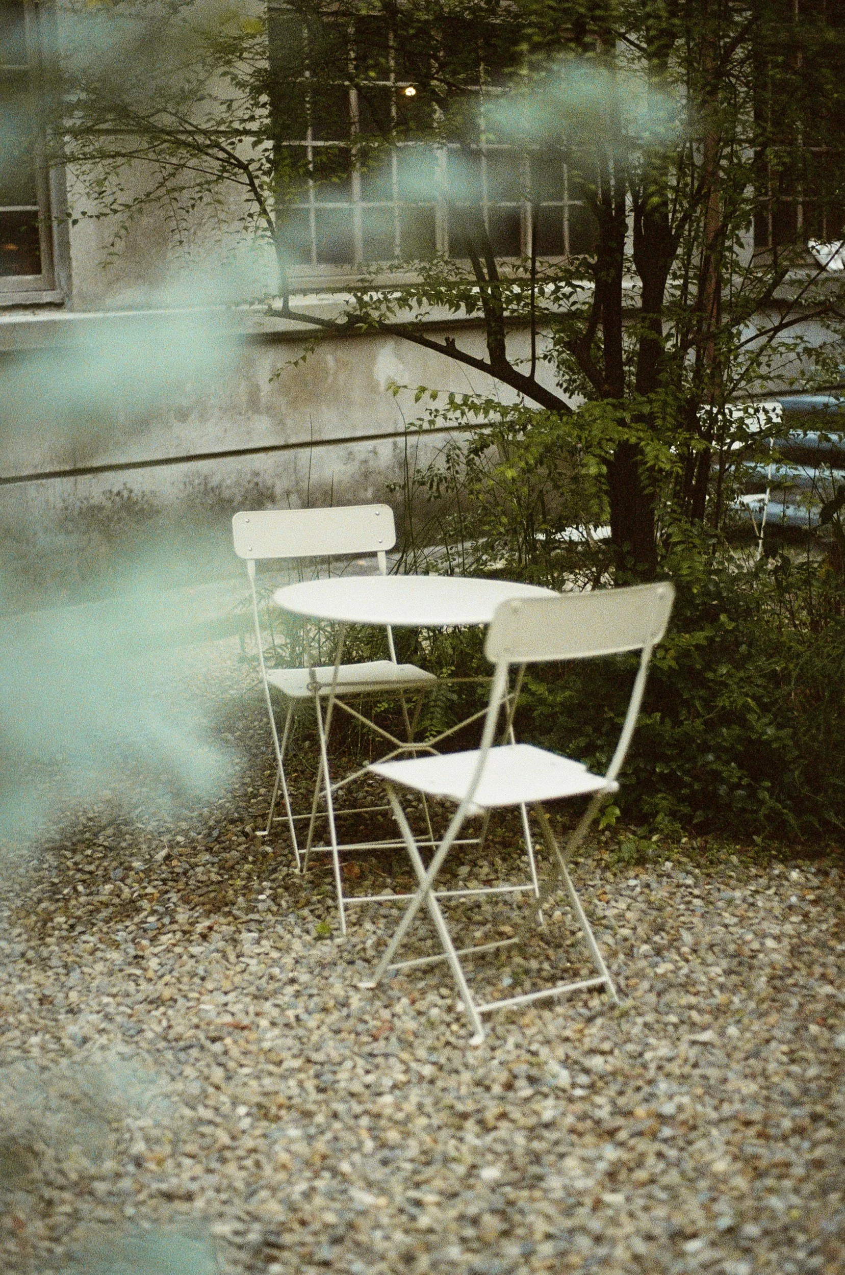 Empty white metal outdoor table with two matching white chairs on gravel ground, surrounded by greenery and trees, in front of an old building with large windows.