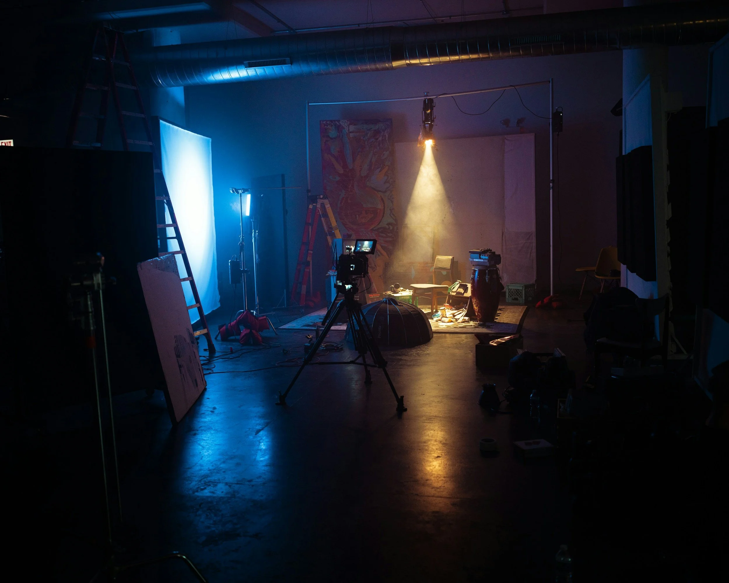 A dimly lit indoor studio setup with various lighting equipment, a camera on a tripod pointing toward a small artful setup with chairs, a sculpture, and a backdrop, indicating a filmmaking or photography shoot in progress.