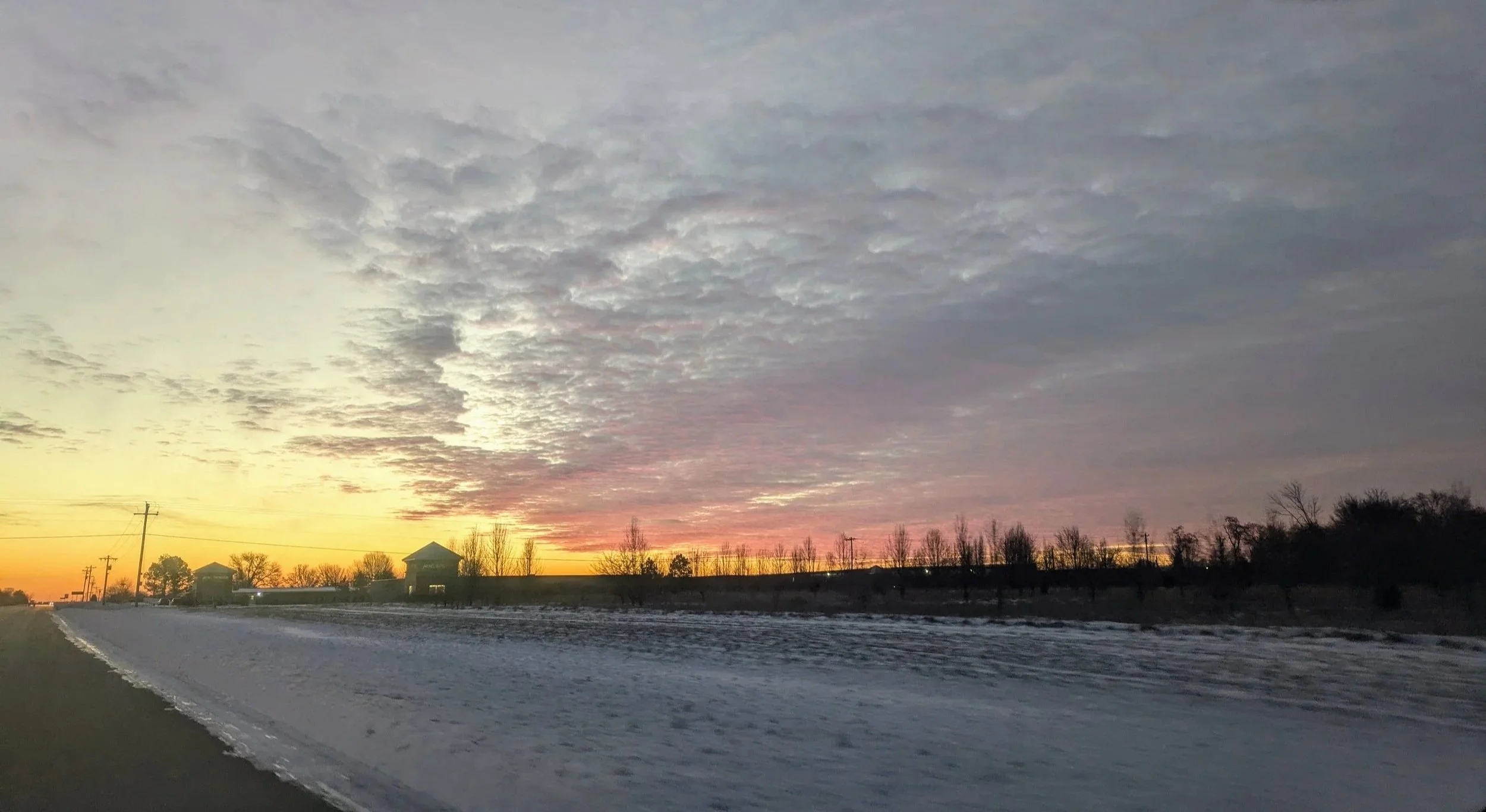 A winter sunset over a rural landscape with a snow-covered road, trees, and a few small buildings under cloudy skies.