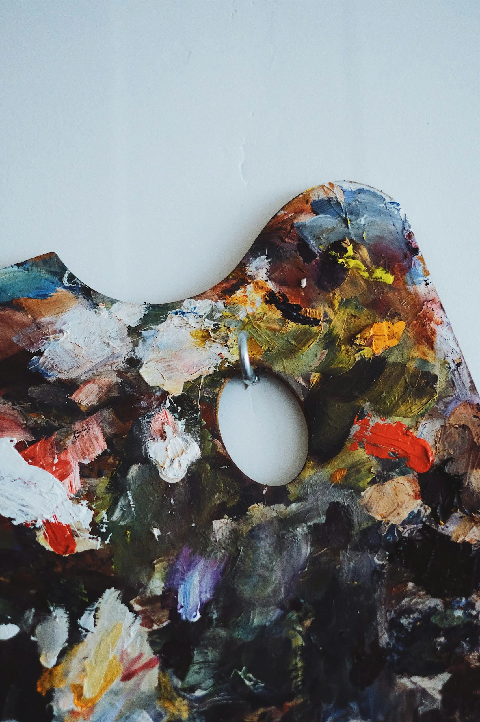 Close-up of an artist's wooden palette with colorful paint strokes, hanging on a hook on a plain white wall.