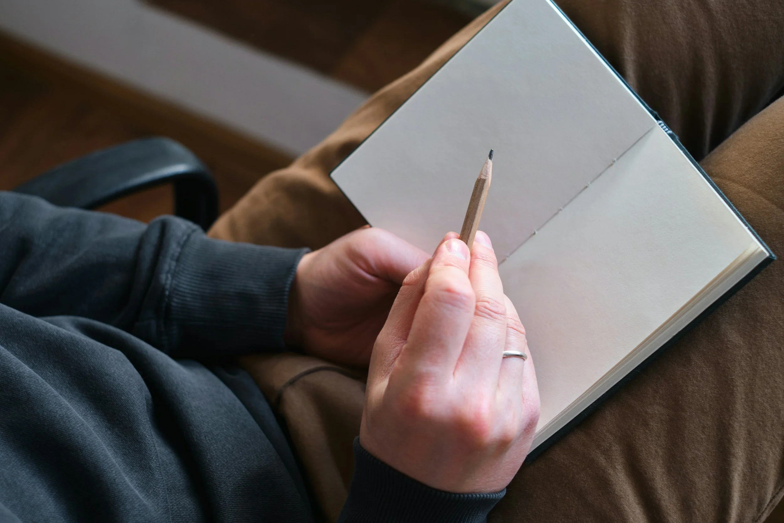 Person holding a notebook and pencil, sitting on a brown couch with a black armrest, writing or drawing in the notebook.