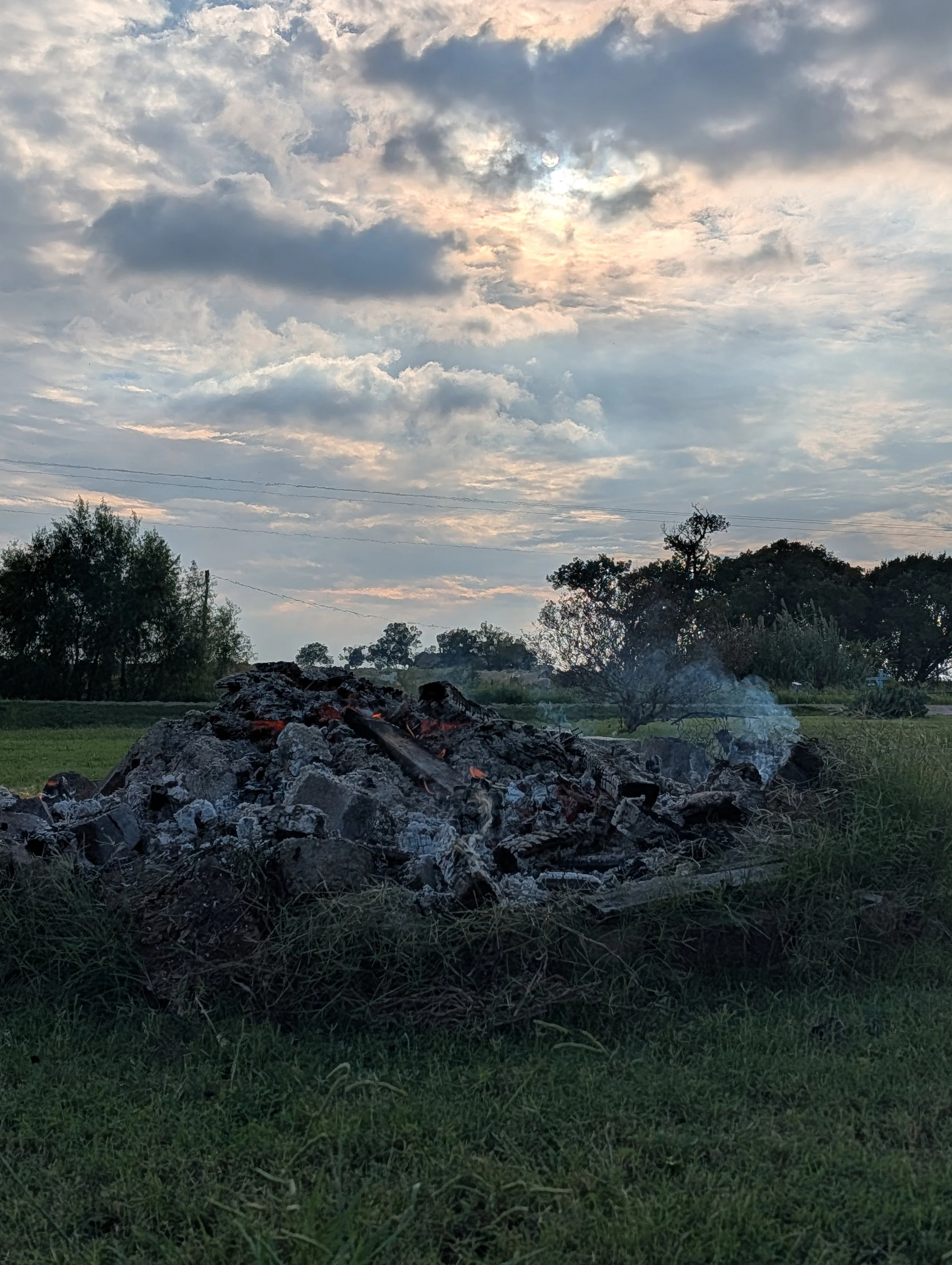 A pile of burning trash with smoke rising, set against a cloudy sky at sunset or early evening, with trees and grassy fields in the background.