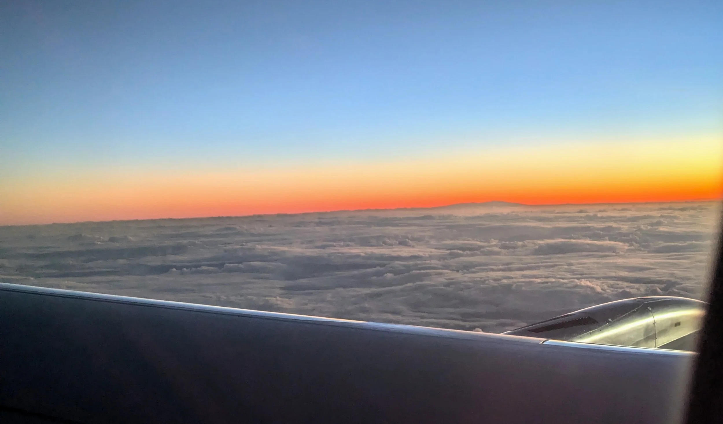View from an airplane window showing a colorful sunset over a sea of clouds with a plane wing visible.
