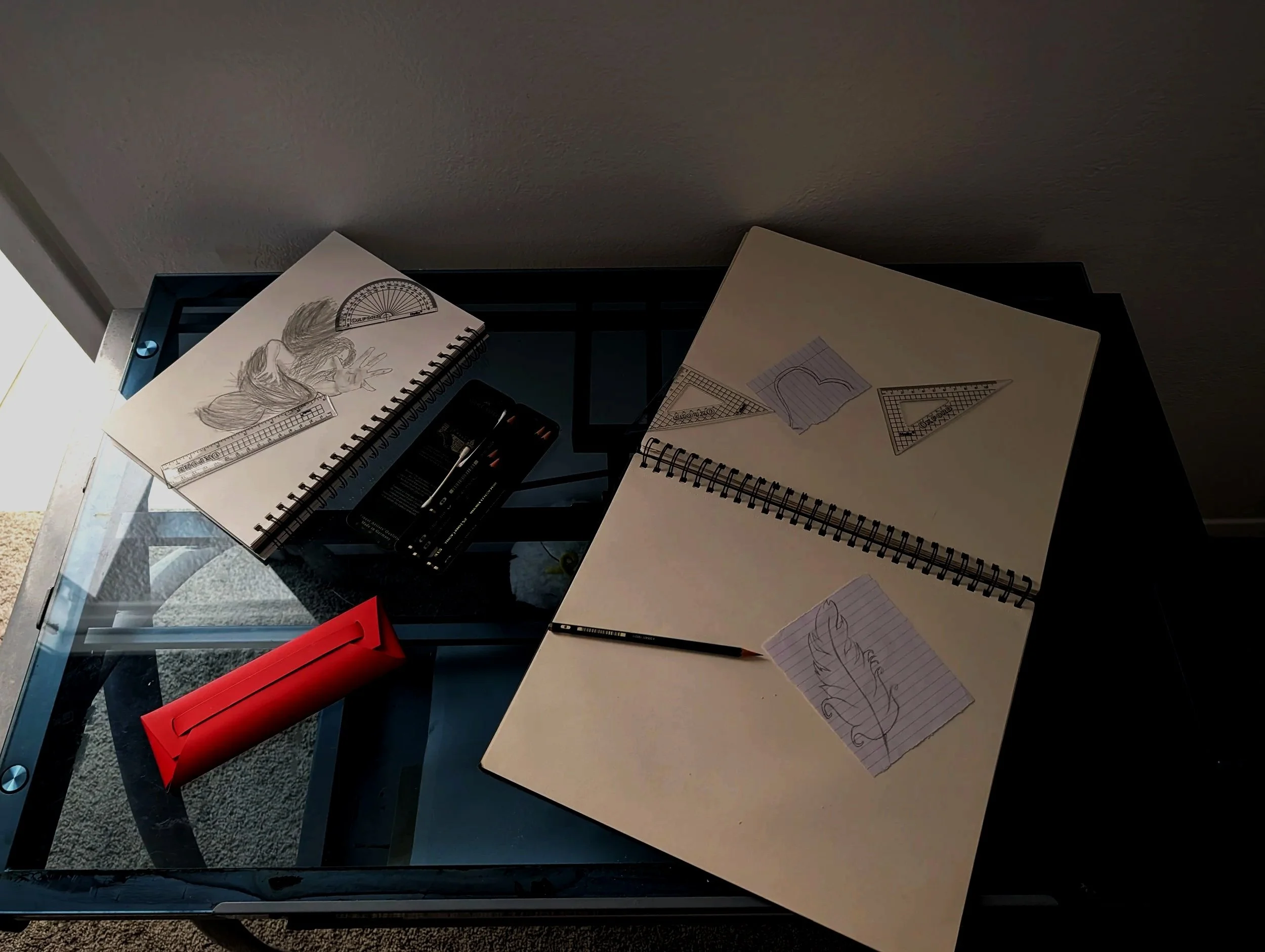 An overhead view of a glass table with drawing supplies, sketchbooks, and paper. The table contains a sketch of a person, geometric rulers, a red pencil case, a black pencil, and sheets of paper with sketches.