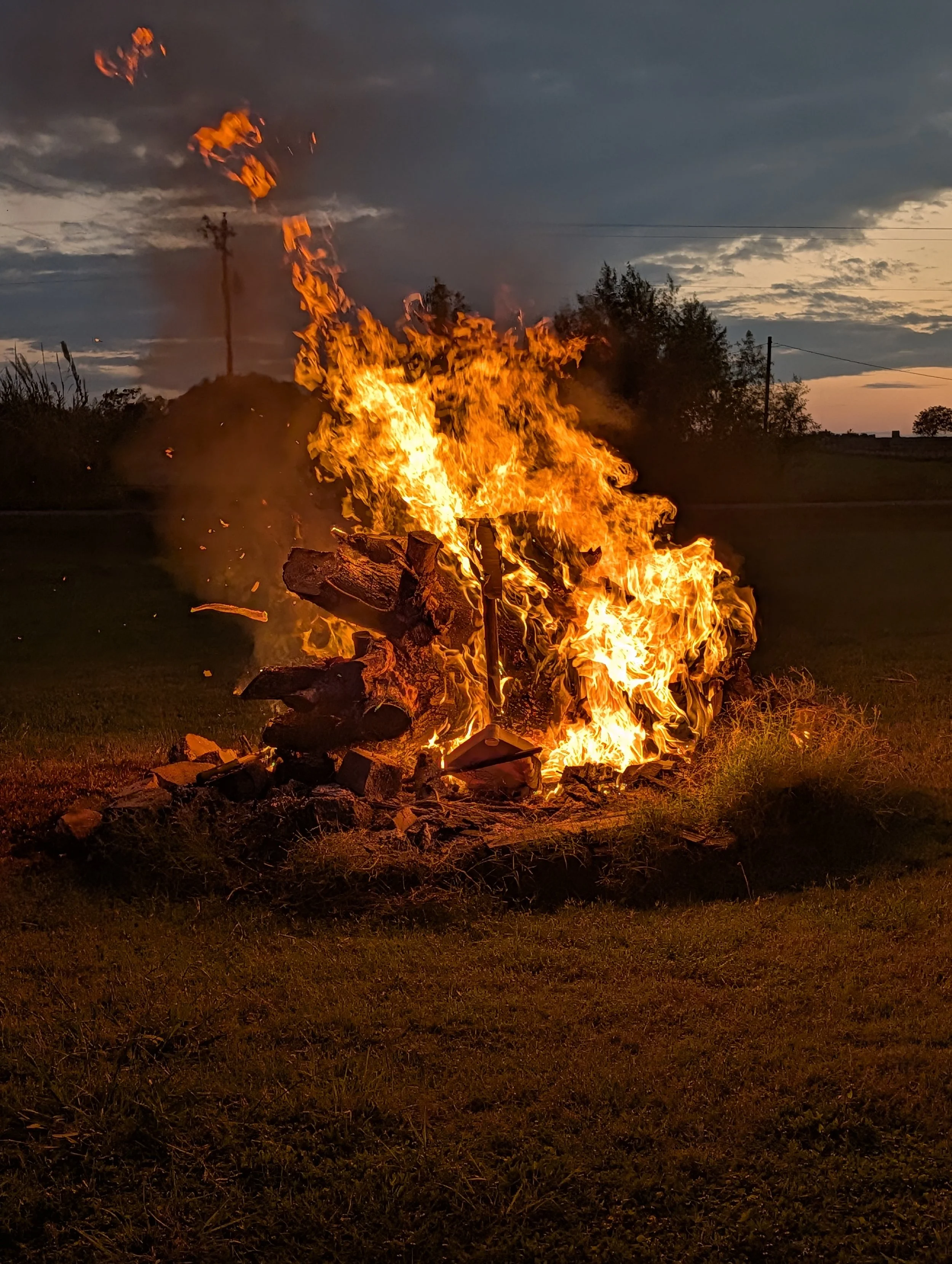 A large outdoor fire burning a pile of wooden debris on a grassy area during dusk.