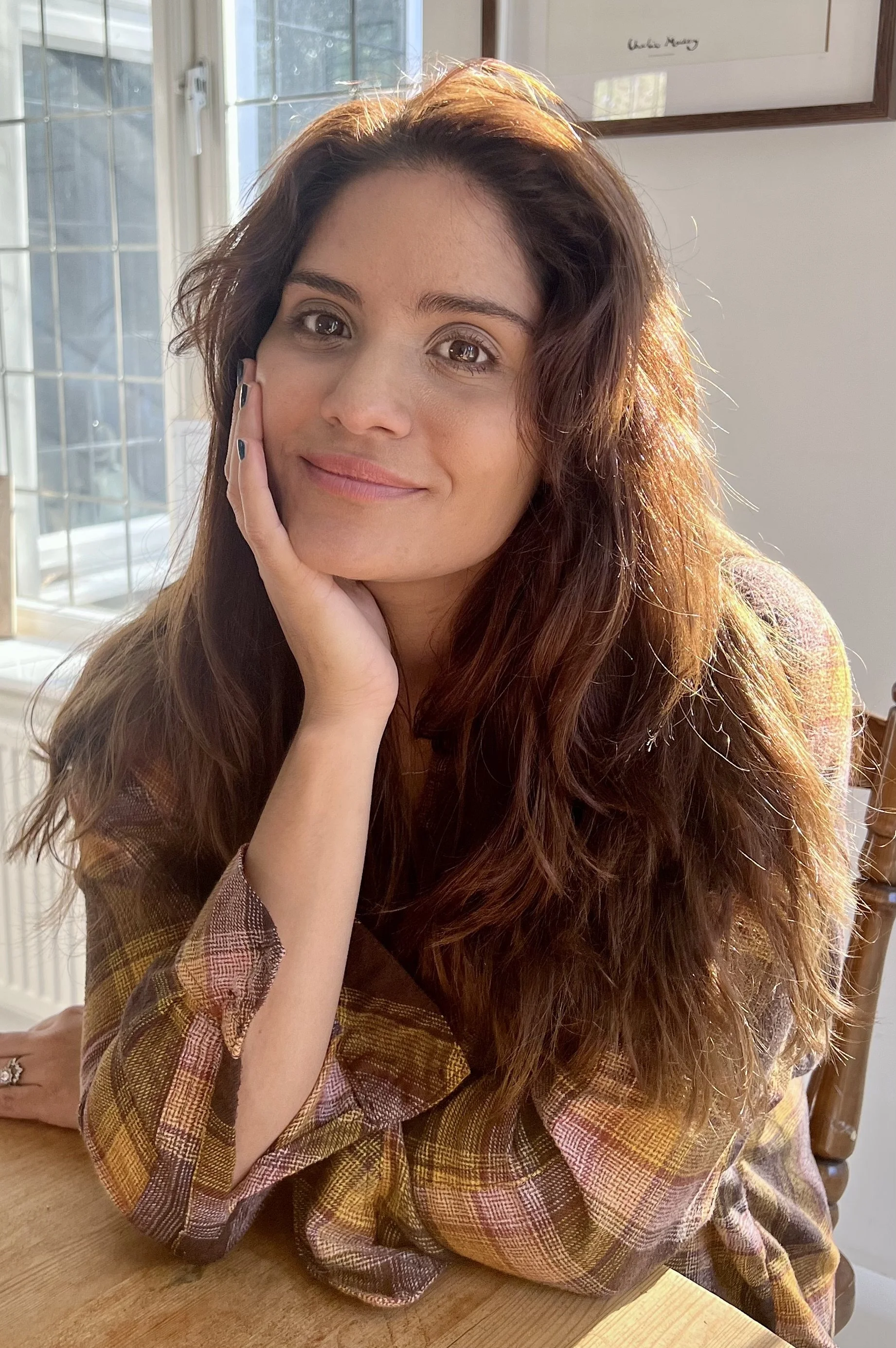 A woman with long, wavy brown hair sitting at a wooden table, resting her head on her hand, with sunlight illuminating her face and hair, and a window with a patterned glass in the background.