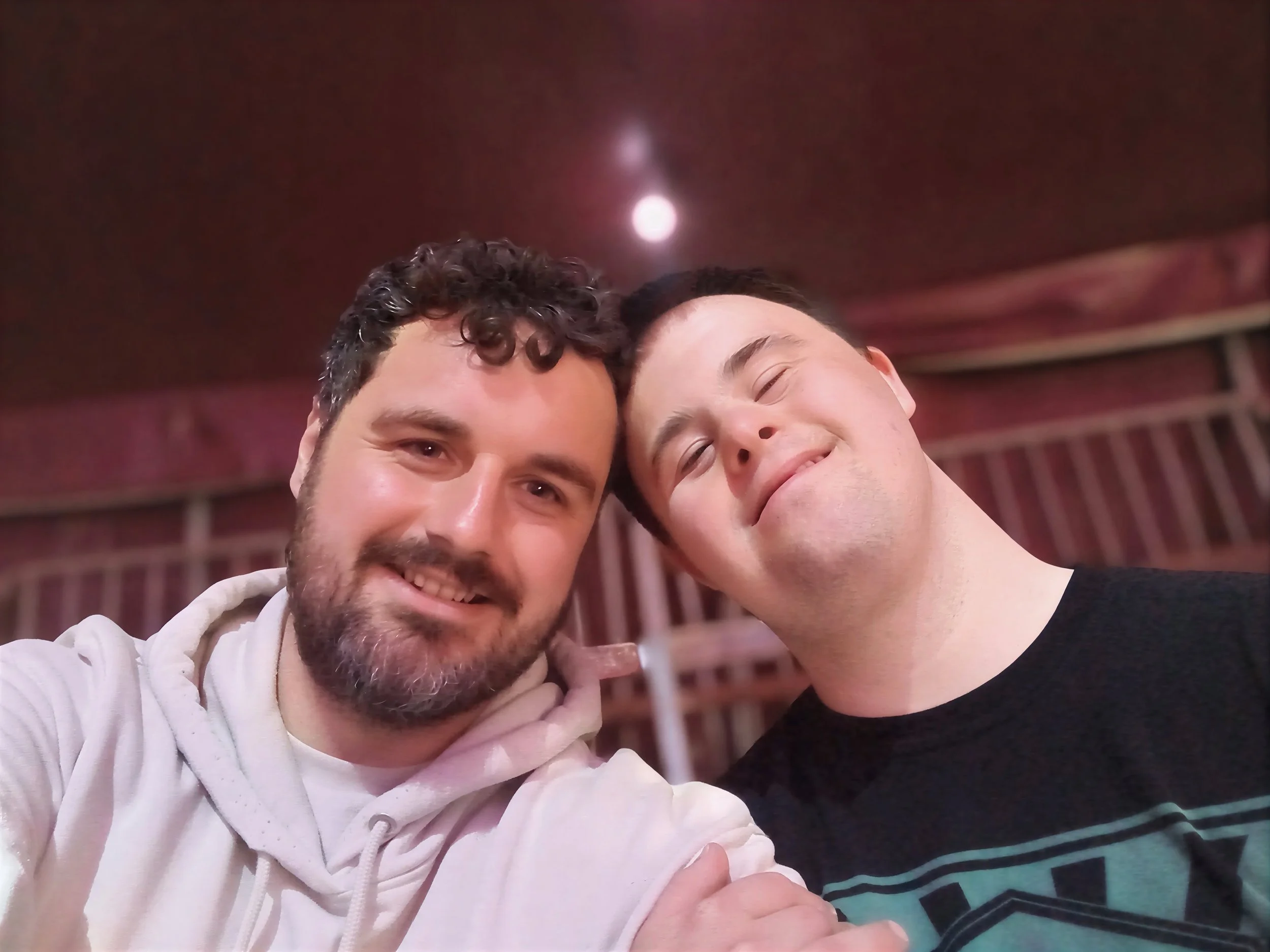 Two men are close together, smiling and posing for a selfie. One man has curly dark hair and a beard, wearing a white hoodie. The other man has short dark hair, is clean-shaven, and is wearing a black shirt with green and purple stripes. They appear to be in a cozy indoor setting with a red ceiling and wooden wall accents.