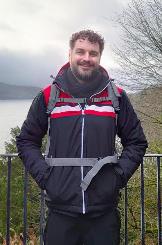 Man with curly hair and beard smiling outdoors, wearing a black jacket with red and white stripes, carrying a backpack, standing in front of a railing with trees and a body of water in the background.