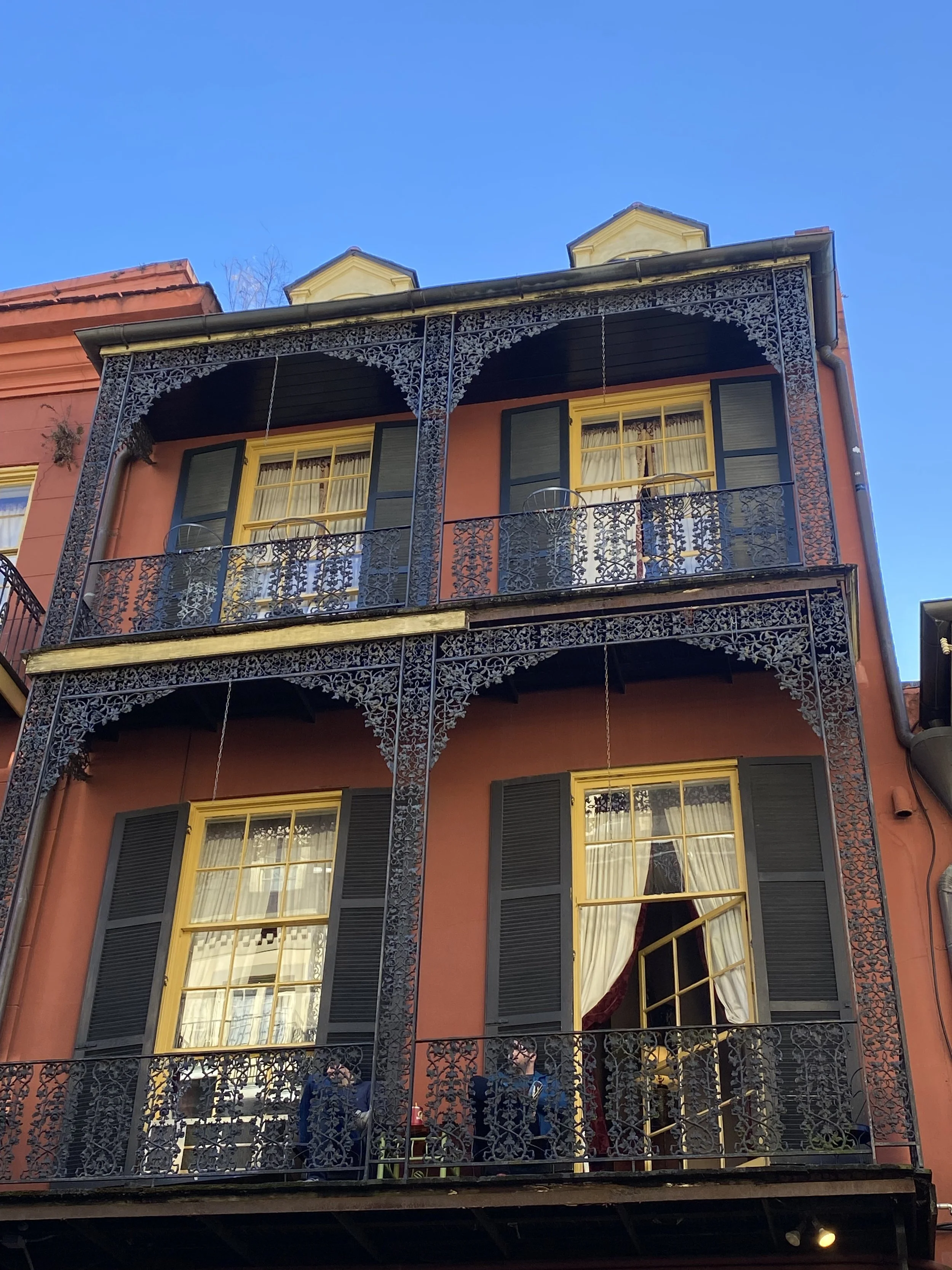 Three-story building with decorative wrought iron balconies, yellow window frames, and dark shutters. Two people are seated on the bottom balcony, and the upper balconies are empty. The building has a reddish facade and a clear blue sky above.