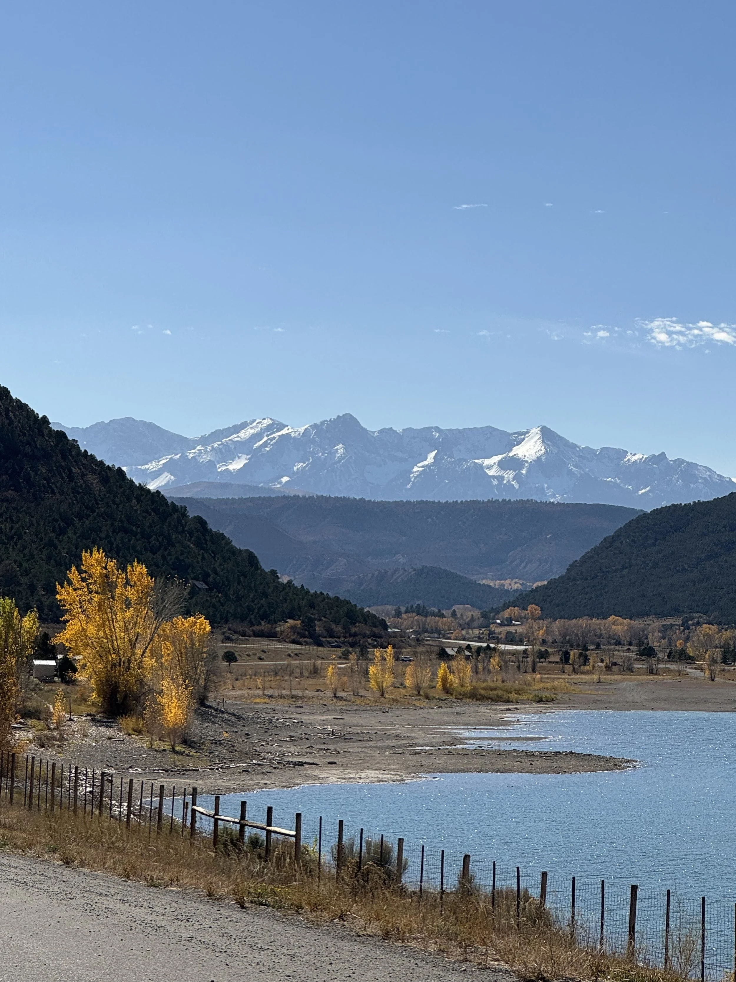 Scenic view of a lake with a gravel shoreline, surrounded by trees with yellow autumn leaves, green mountains, and snow-capped peaks in the distance under a clear blue sky.