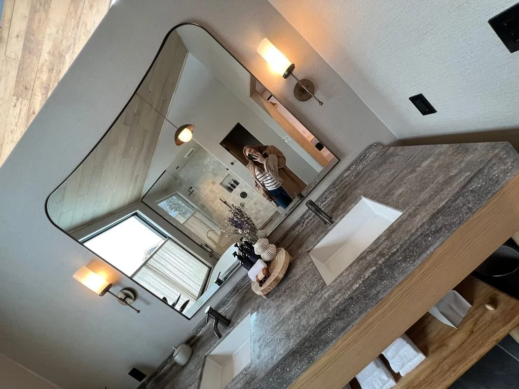 A bathroom vanity with a gray granite countertop, two rectangular white sinks, and a large rectangular mirror above. The reflection shows a woman taking a photo in a mirror. The bathroom features wall-mounted light fixtures, a window, and decorative items on the countertop.