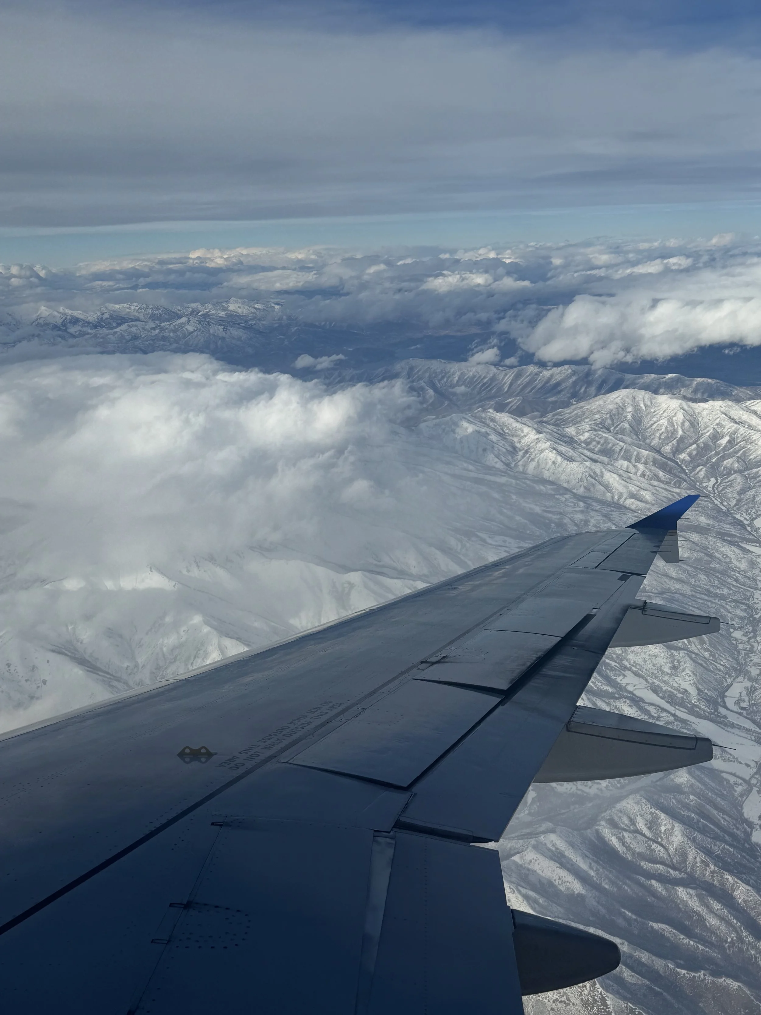 View from an airplane window showing a snow-covered mountain range below with clouds covering parts of the landscape.