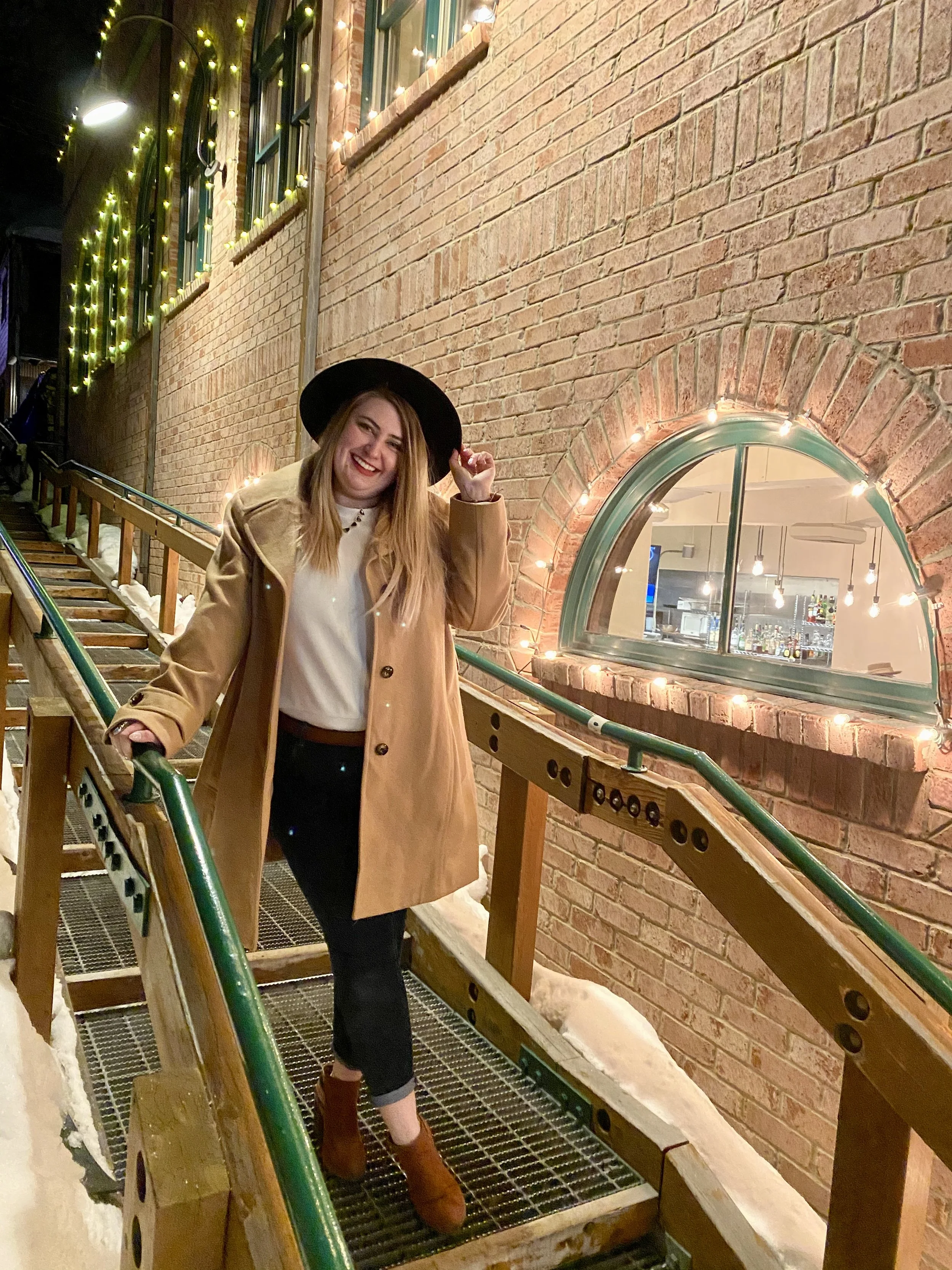 A young woman with long blonde hair wearing a black hat, tan coat, white sweater, black pants, and brown ankle boots, smiling and standing on a metal and wood staircase with green railings, next to a brick building decorated with string lights around a large arched window at night.