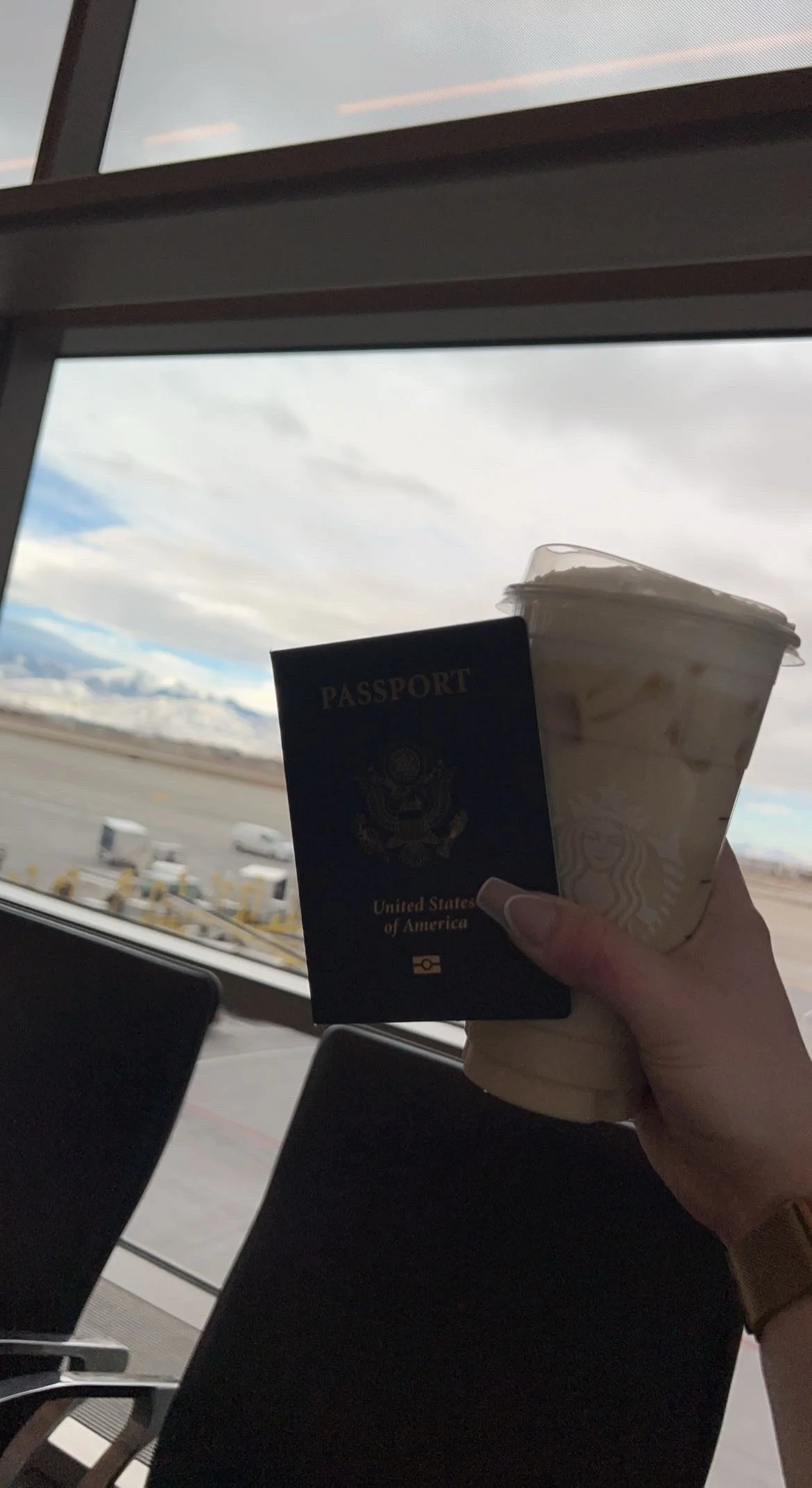 A hand holding a U.S. passport and a Starbucks iced coffee in an airport terminal with view of the tarmac and cloudy sky outside.