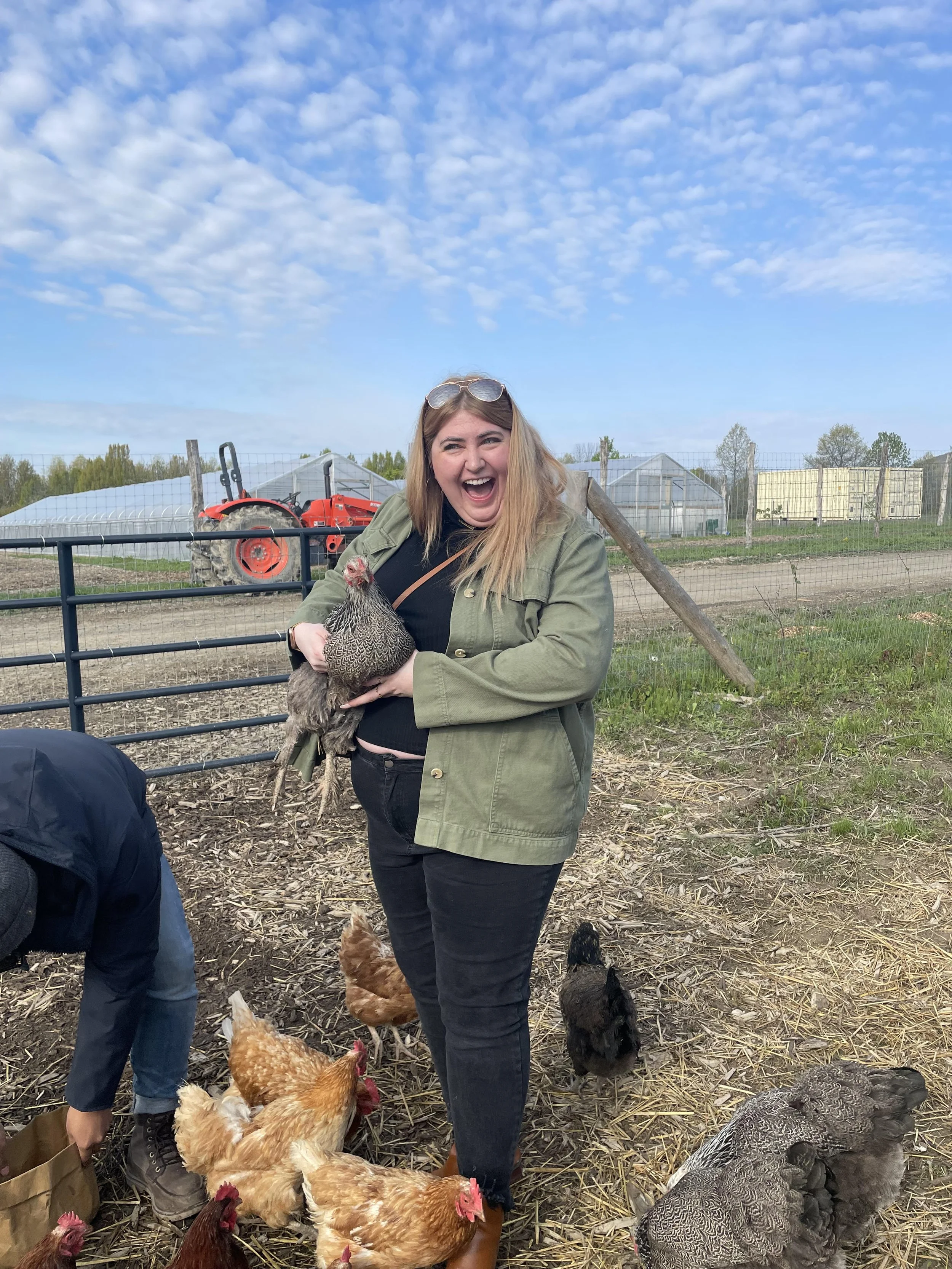 A woman with long blonde hair, wearing a green jacket and black pants, holding a chicken in an outdoor farm setting with some chickens at her feet. She is smiling and laughing, with blue sky and farm structures in the background.