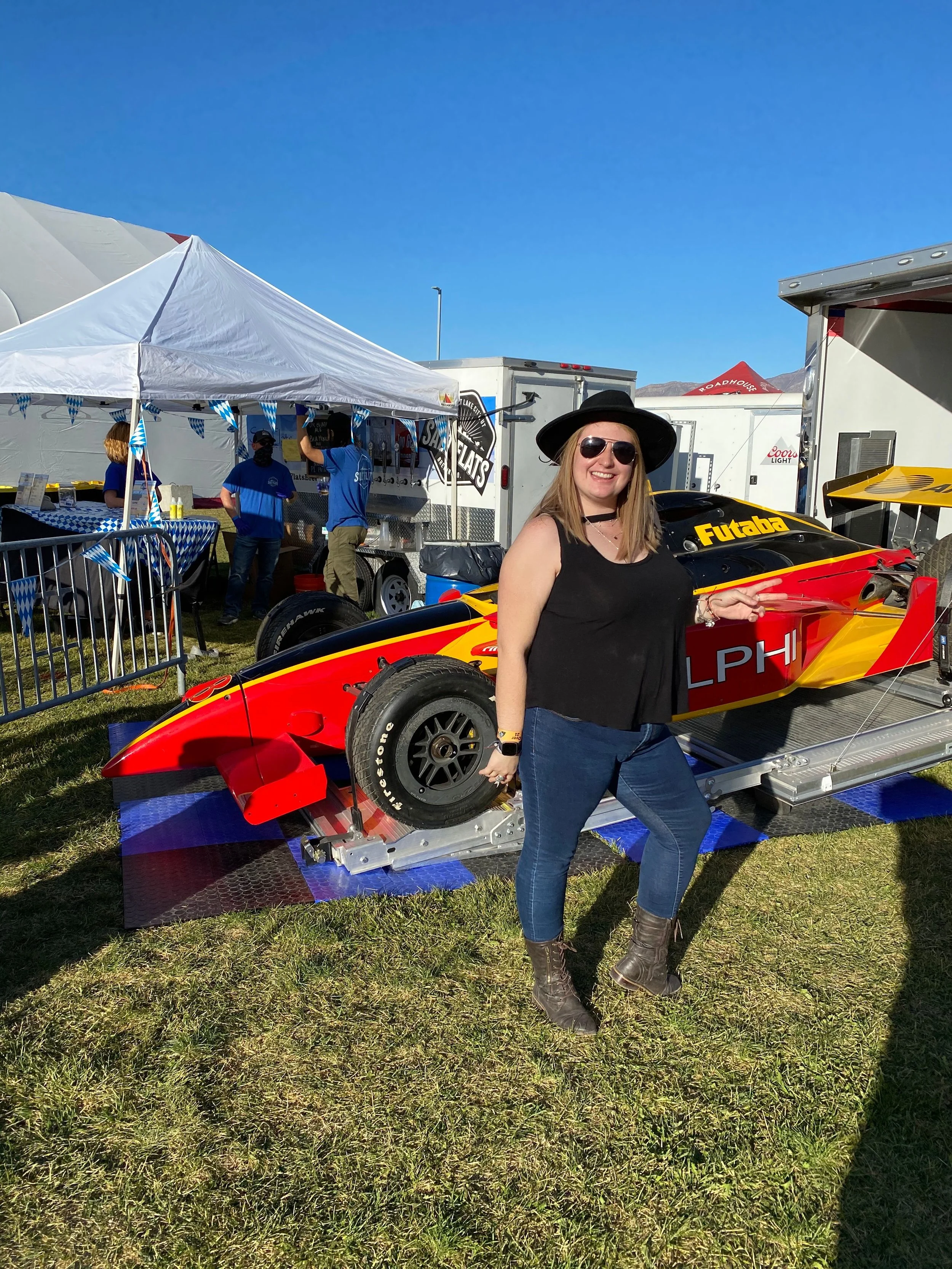 Woman in sunglasses, black hat, and black tank top pointing at a race car on display outside at an event.