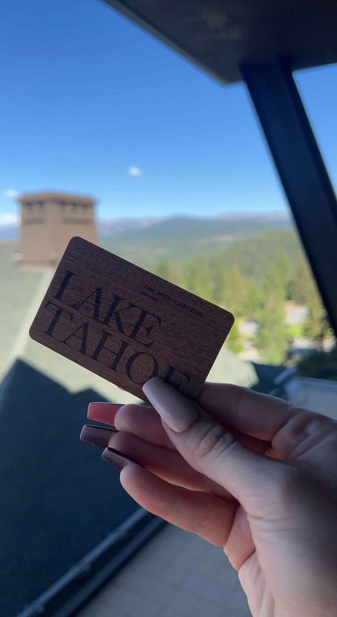 Hand holding a wooden card with the text 'LAKE TAHOE' in front of a mountain landscape with a building and blue sky in the background.