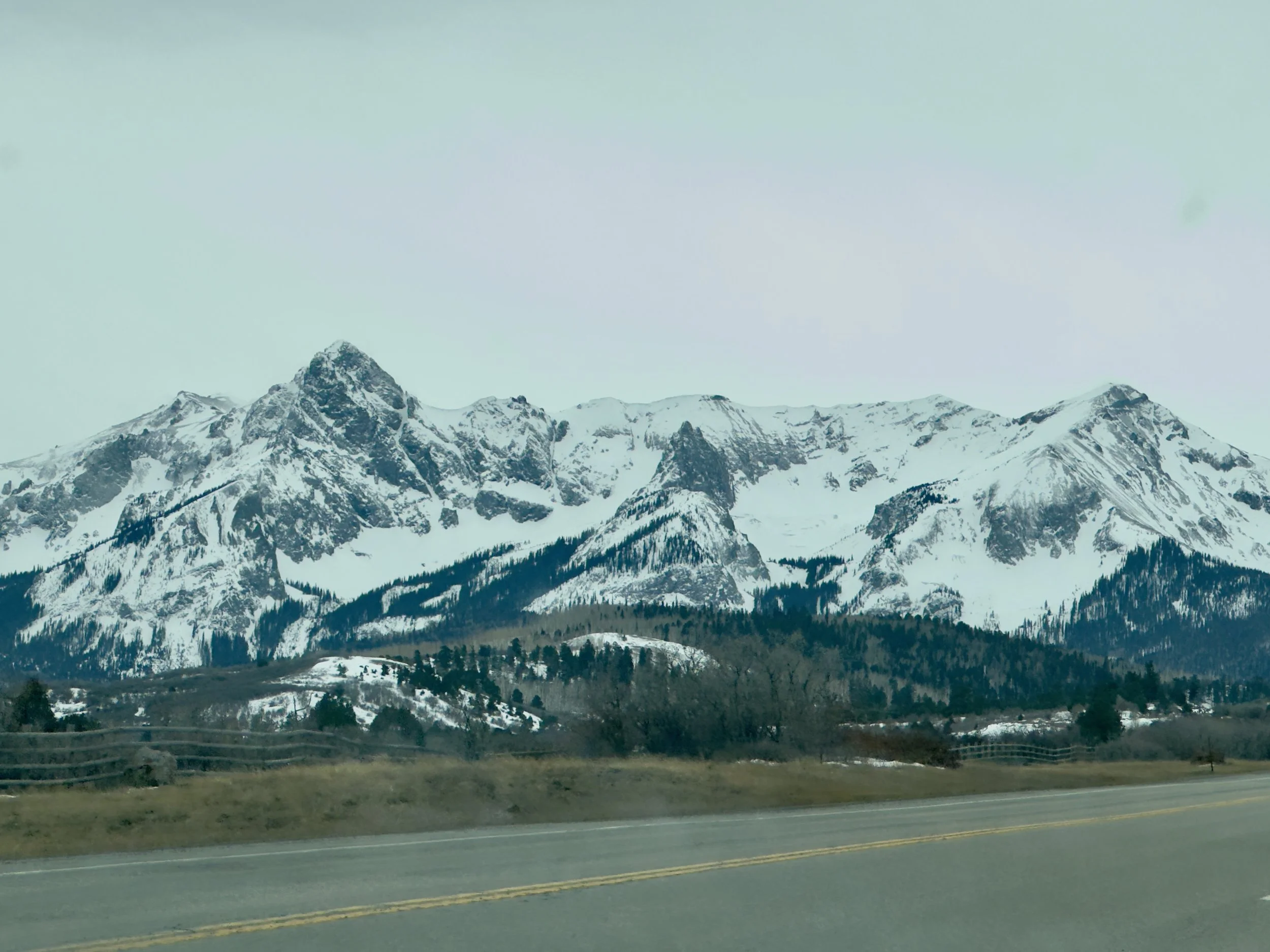 Snow-covered mountain range seen from a roadside view, with a highway in the foreground and a cloudy sky above.
