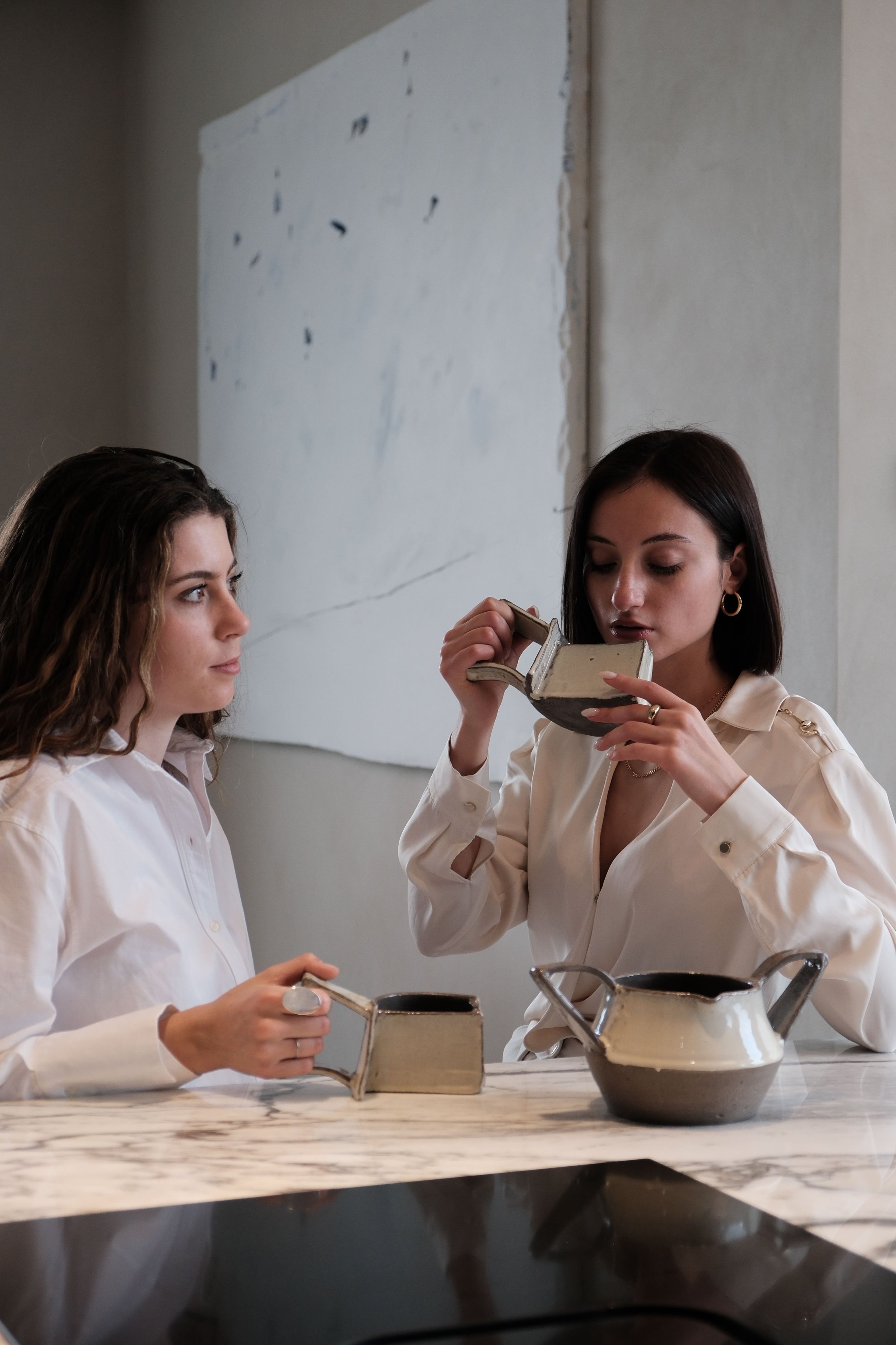 Two women in white shirts preparing chocolate in a kitchen. One woman is holding a small saucepan, and the other woman is inspecting a ceramic bowl. There are pots on a marble countertop and a plain wall in the background.