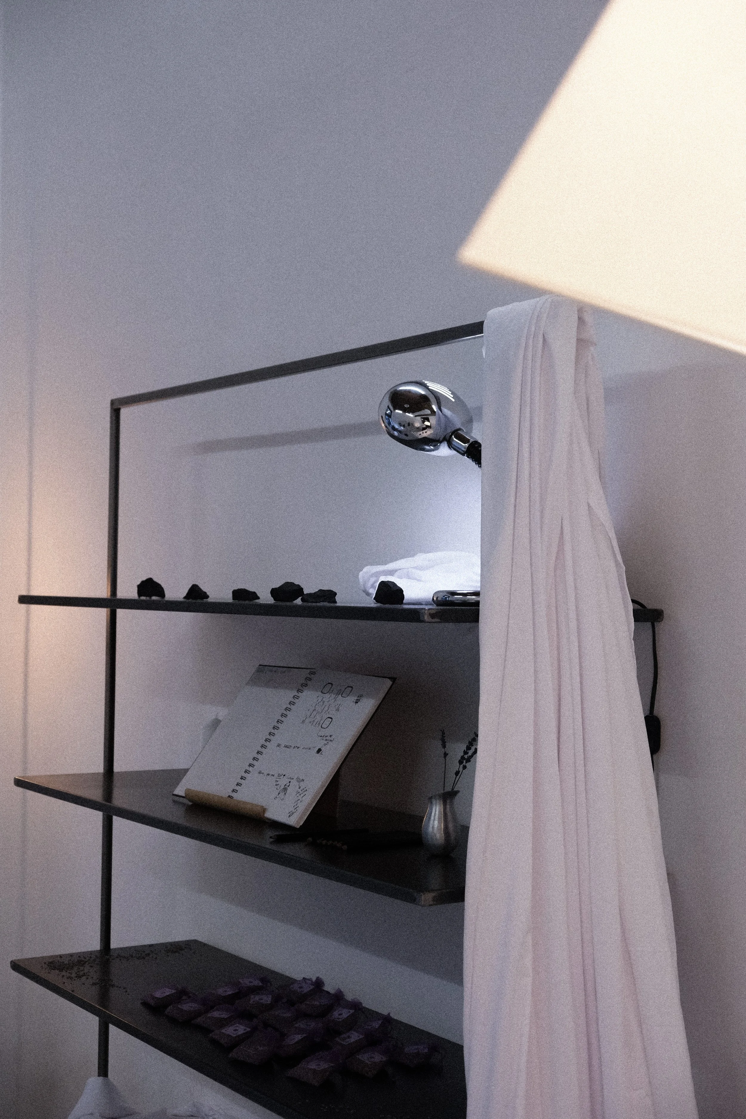 A modern display shelf with four levels. The top level has a reflective security camera or light, with a cloth draped partially covering it. The second level holds a row of small stones or decorative objects, and the third features a large open book 