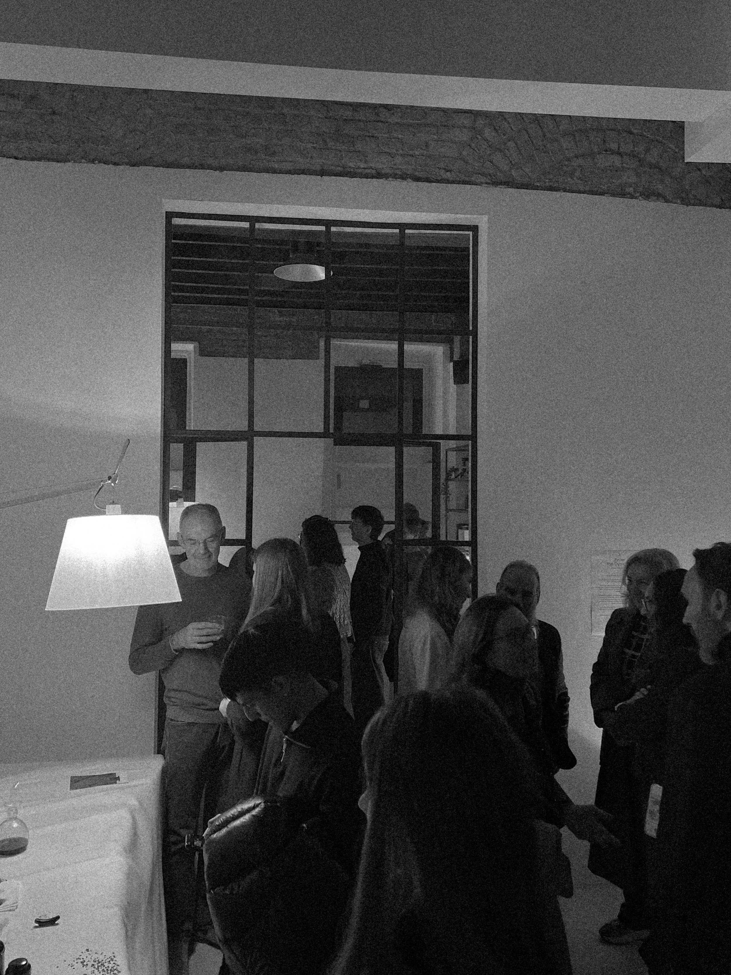 A group of people socializing in a dimly lit room, some holding drinks. There is a white lamp on a table in the foreground, and a decorative wooden beam on the ceiling. The room has a modern style with a large glass window or partition and some artwo